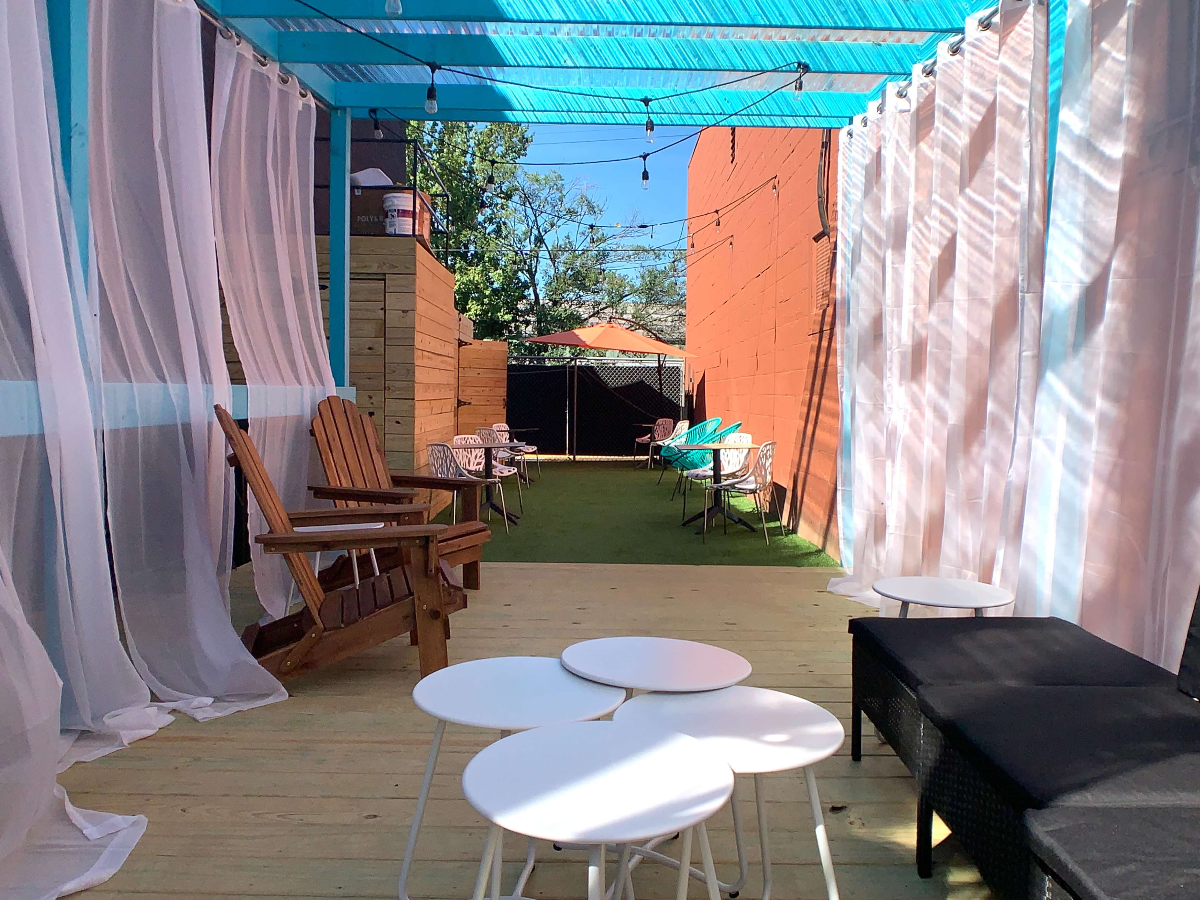 A patio area with a mostly wooden floor, featuring several chairs, tables, and sheer curtains, under a turquoise canopy.