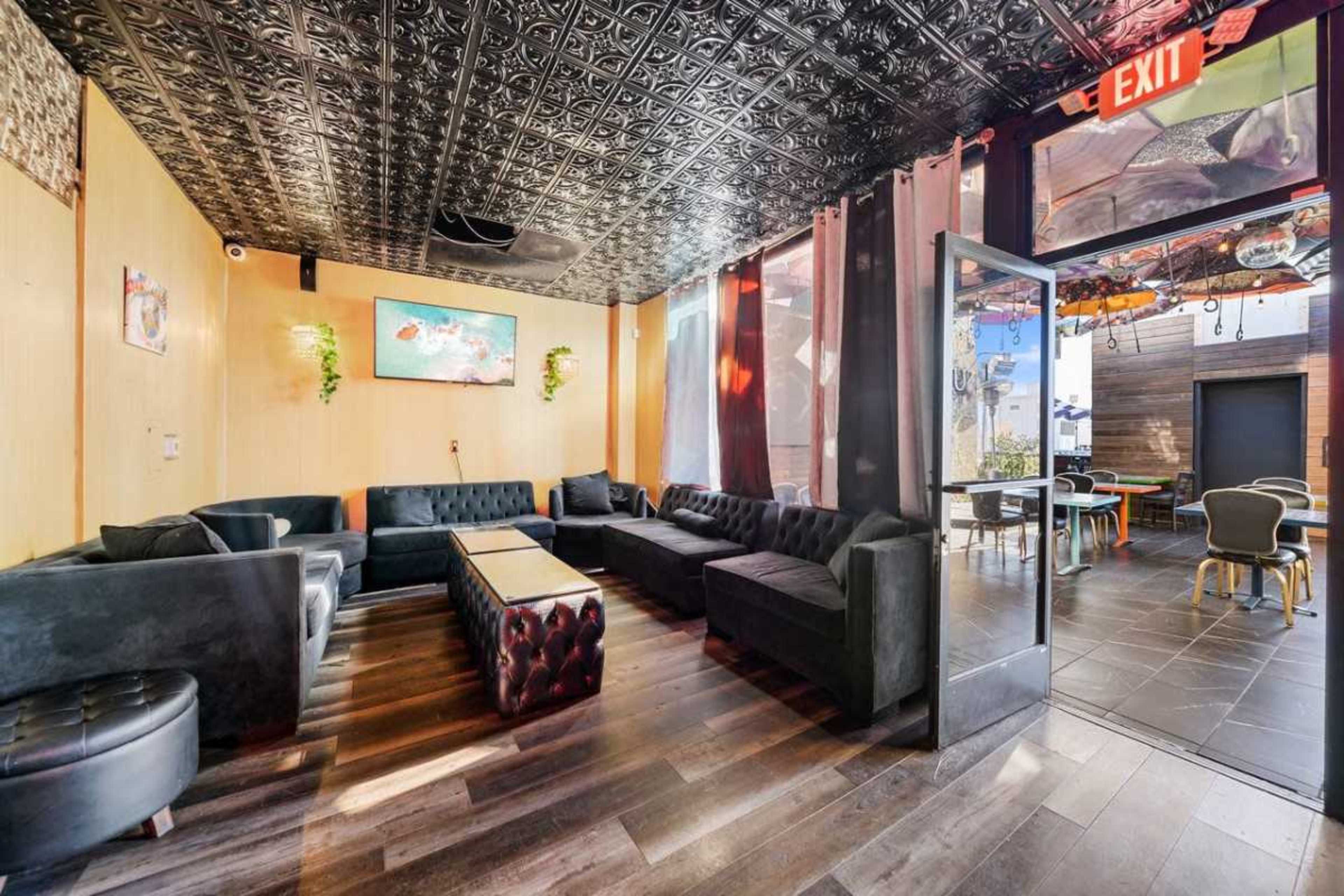 The image shows a lounge area featuring black leather seating arranged around a central wooden table, with large windows and decorative elements on the ceiling.