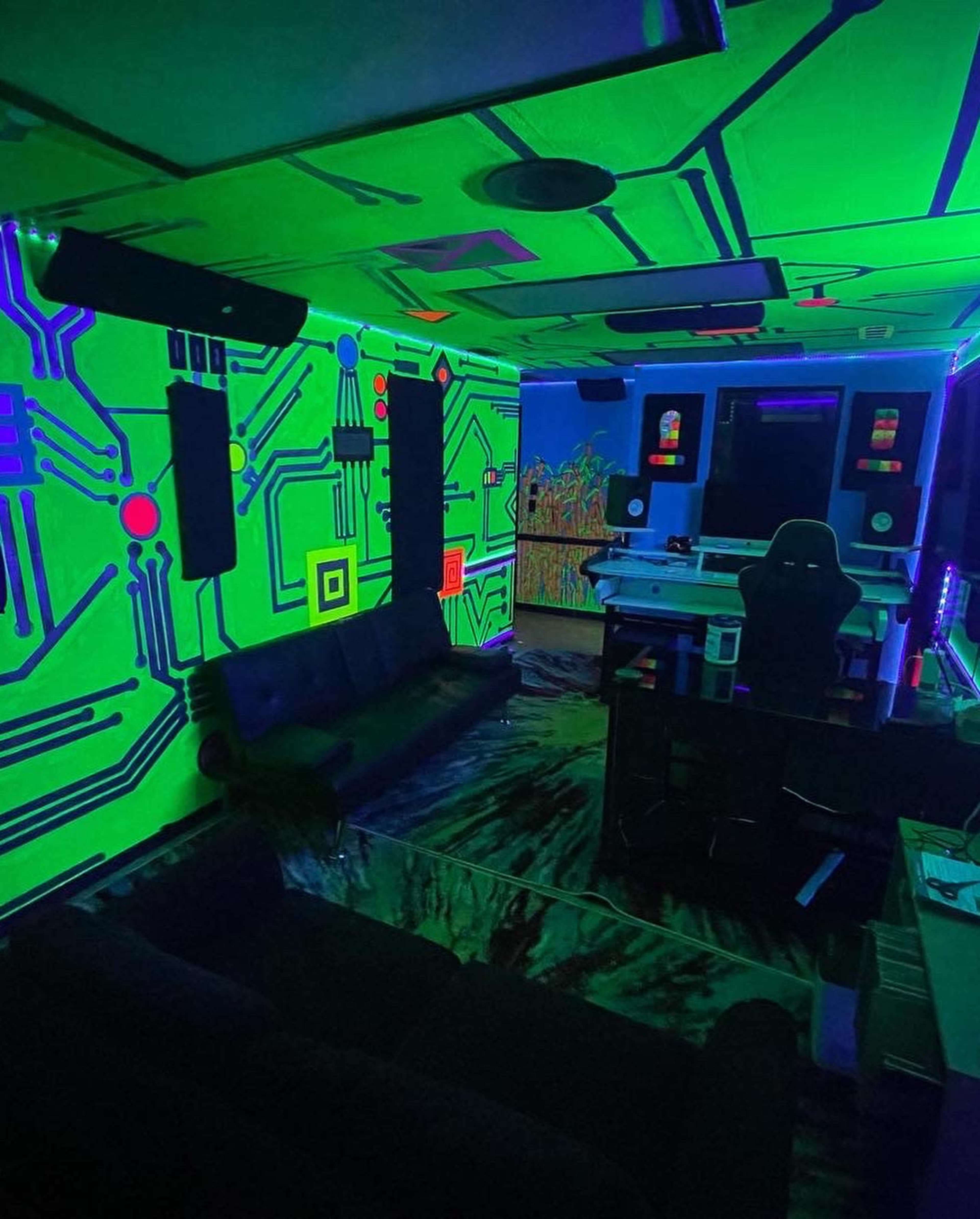 The room features neon circuit board designs on the walls, with black couches and a desk illuminated by colorful lighting.