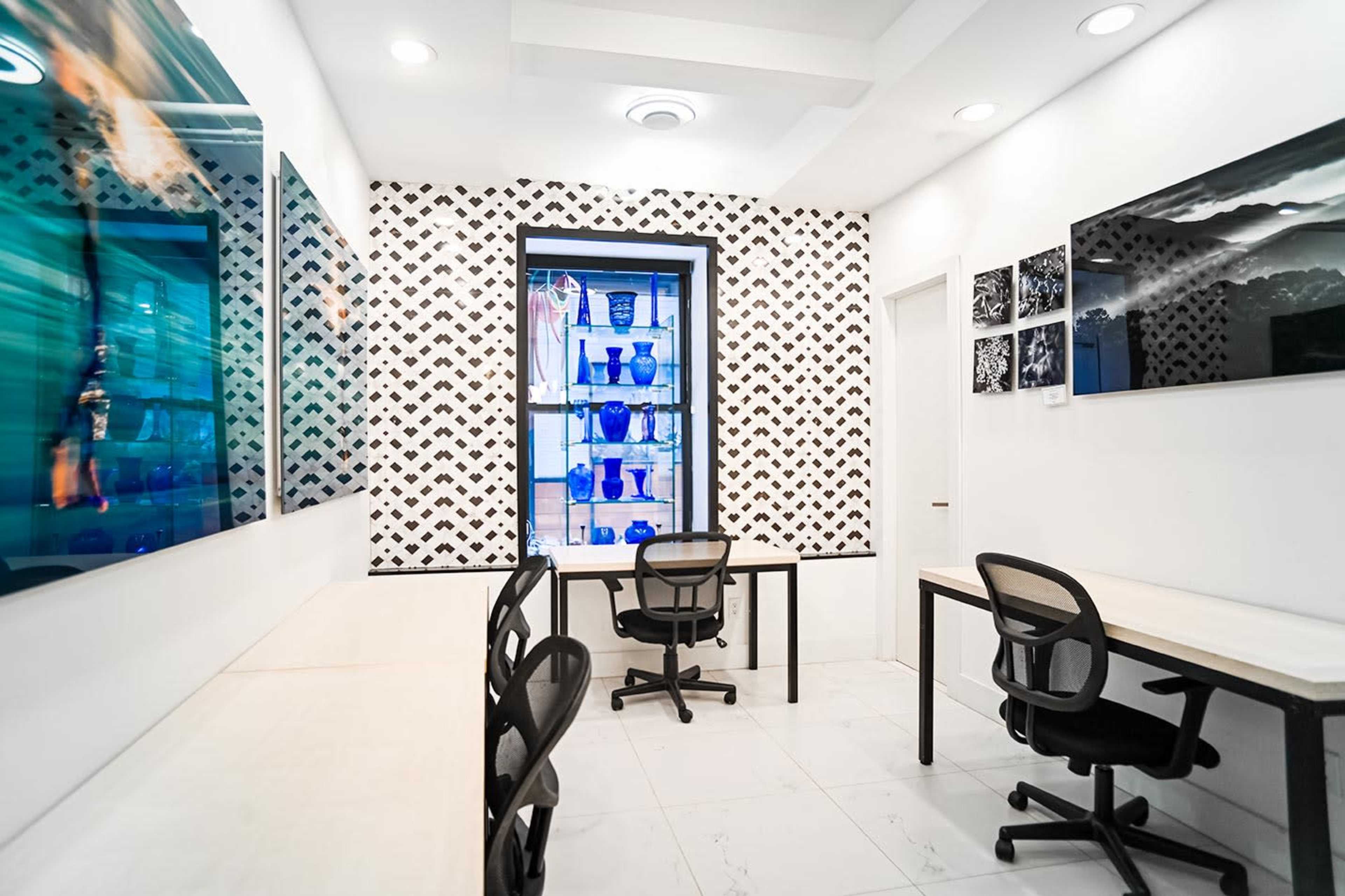 The image shows a modern workspace with two desks and chairs, framed artwork on the walls, and a display case featuring blue glass items.
