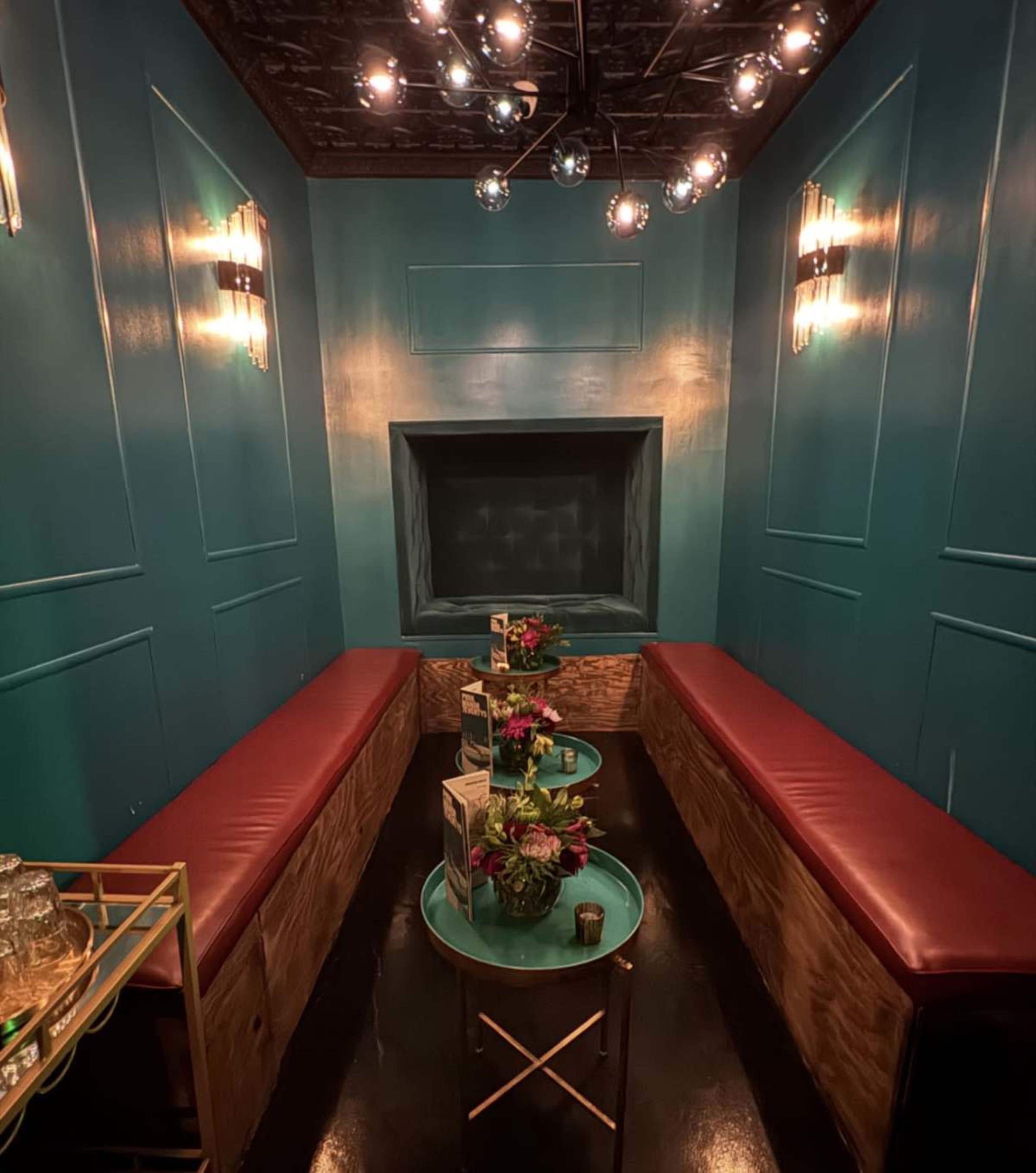 The image shows a small, stylish lounge with teal walls and red leather seating, decorated with plants and a modern lighting fixture.