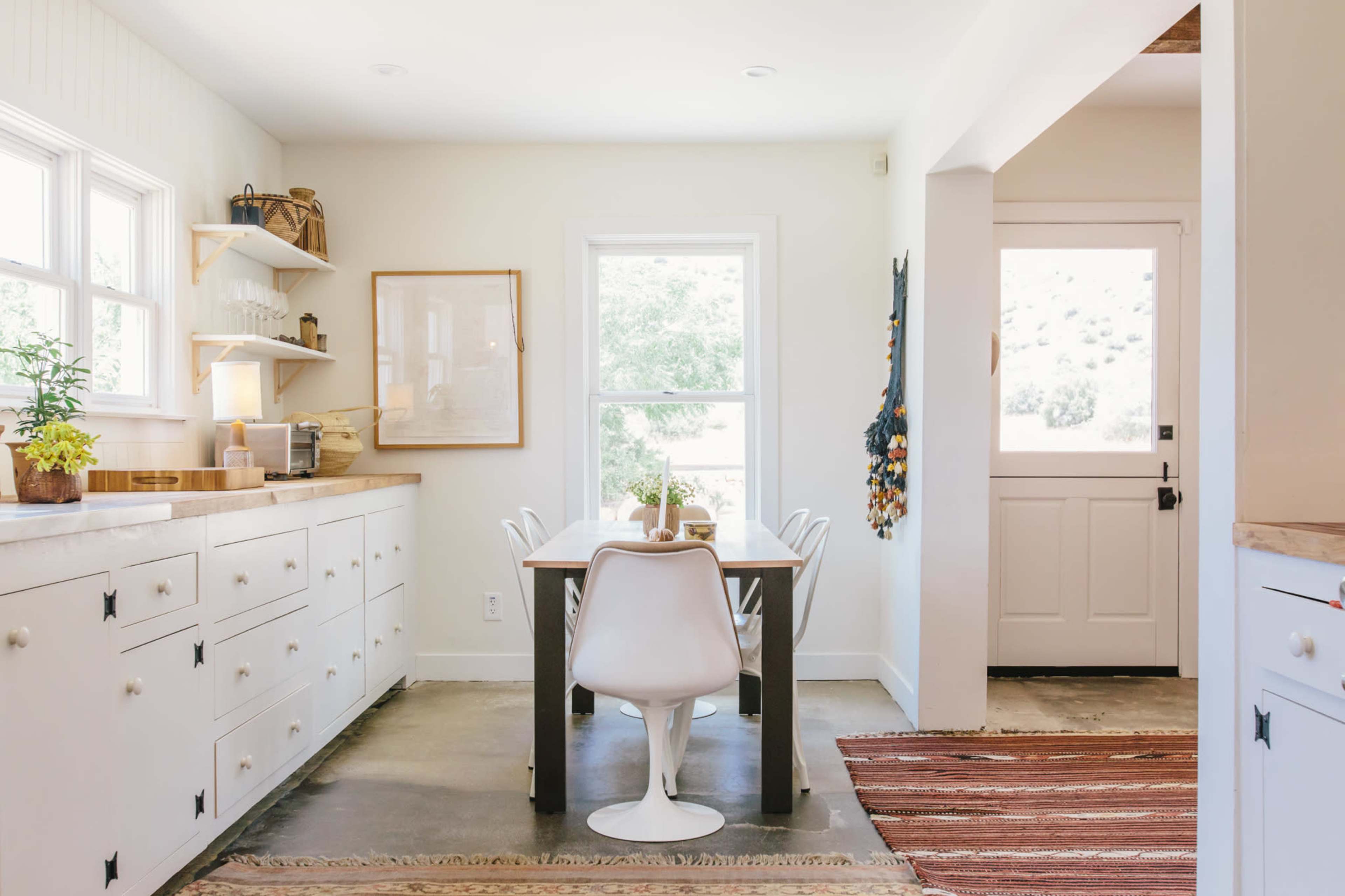 A clean, modern kitchen features a dining table surrounded by chairs, with shelves displaying kitchenware and a door leading to the outside.