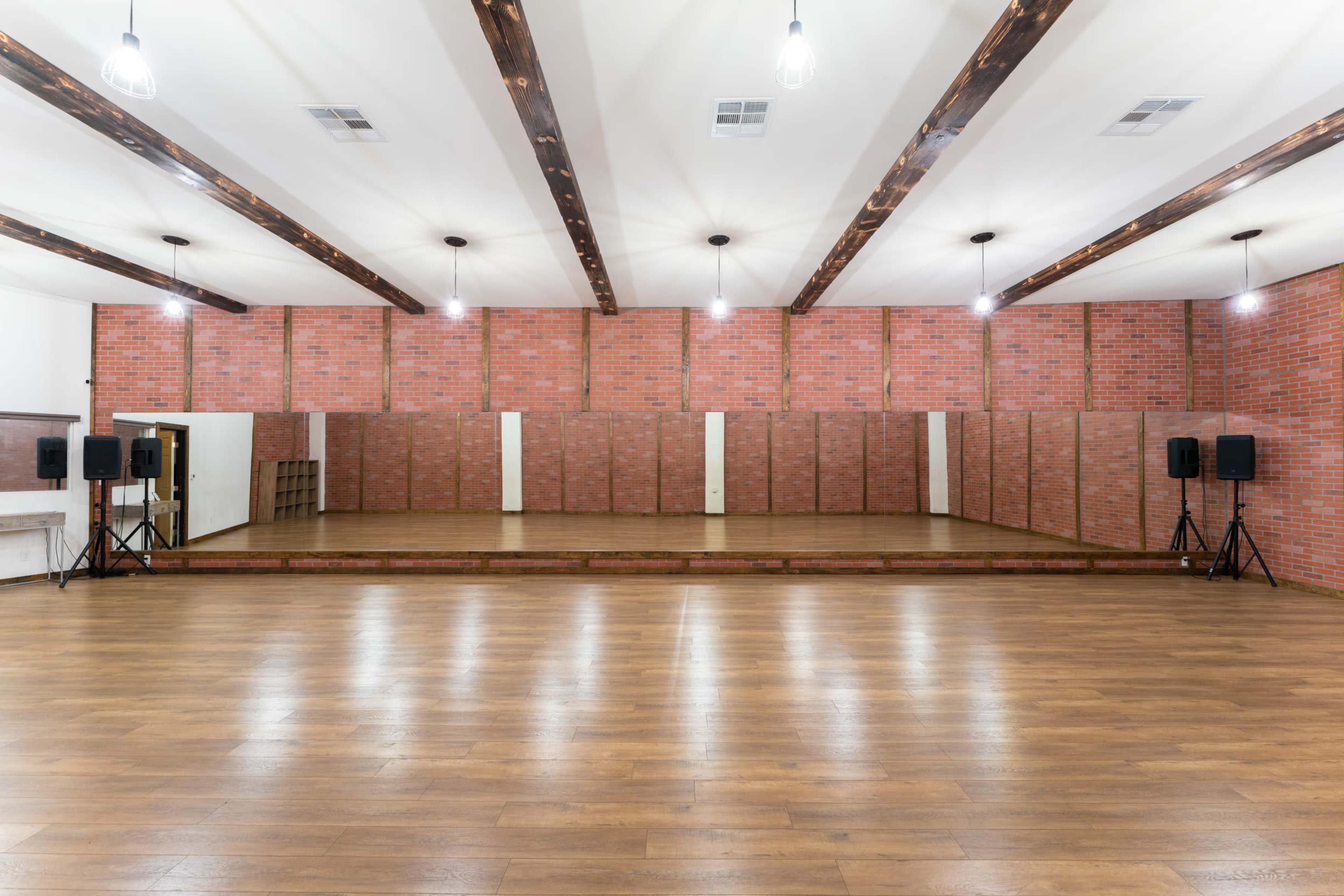 The image shows a spacious dance studio with polished wooden floors, red brick walls, and overhead lights, featuring a mirrored section and portable speakers at both ends.