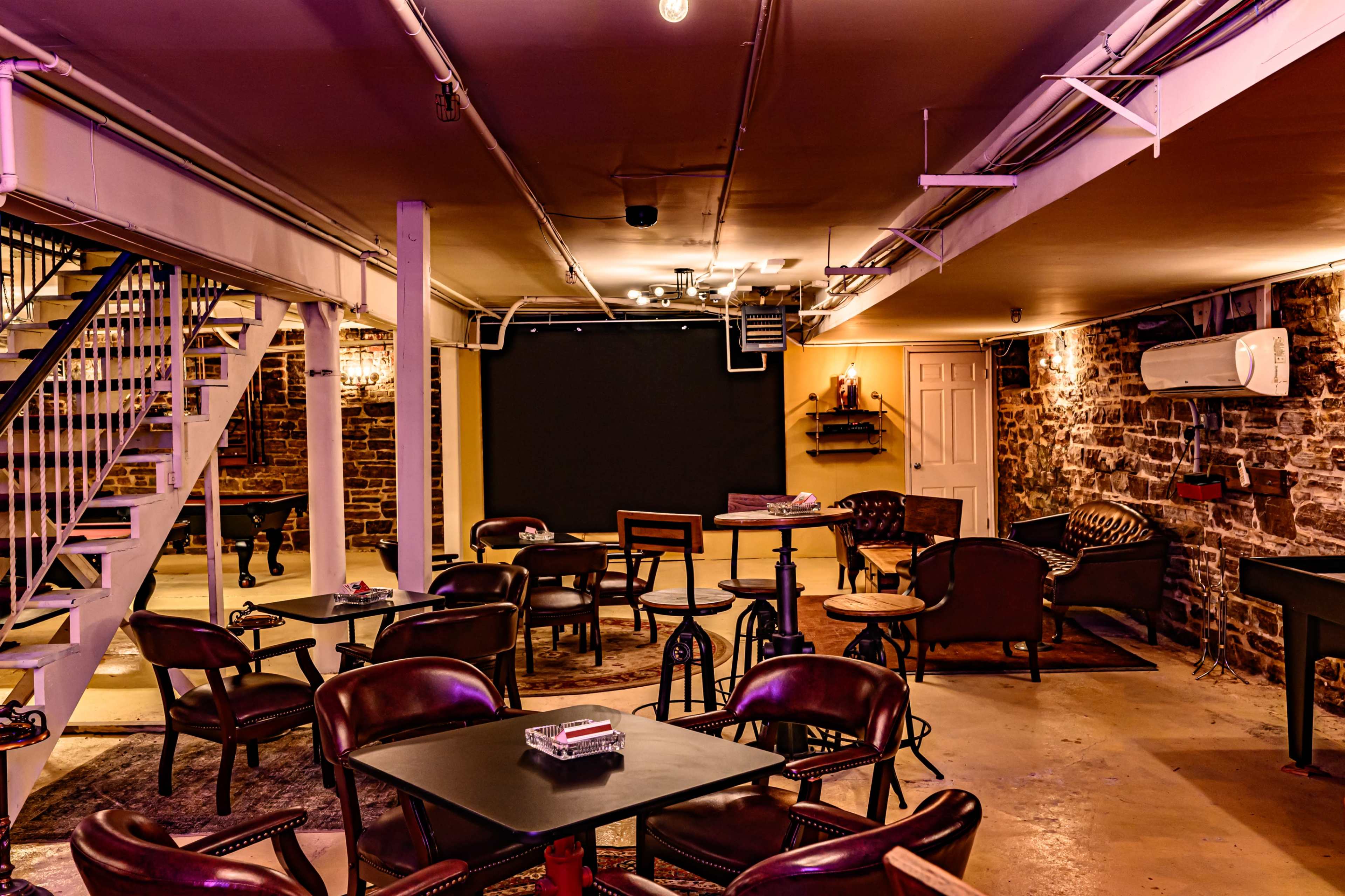 The interior of a dimly lit basement lounge features wooden tables and chairs arranged around a central area, with a staircase leading to another level and a wall adorned with exposed stone.