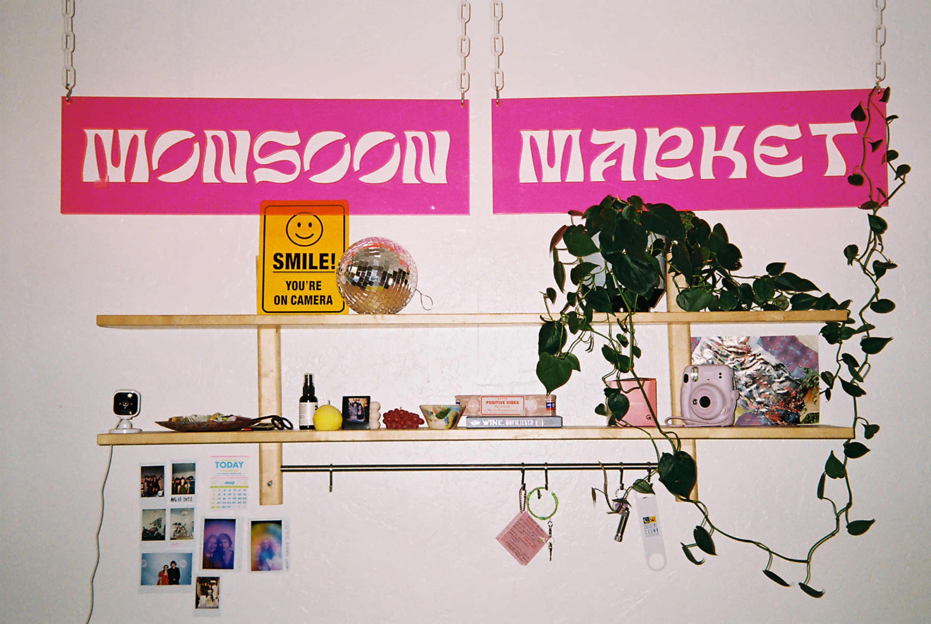 A wall display with two large pink signs labeled "MONSOON" and "MARKET," along with a wooden shelf holding various decorative items and plants.