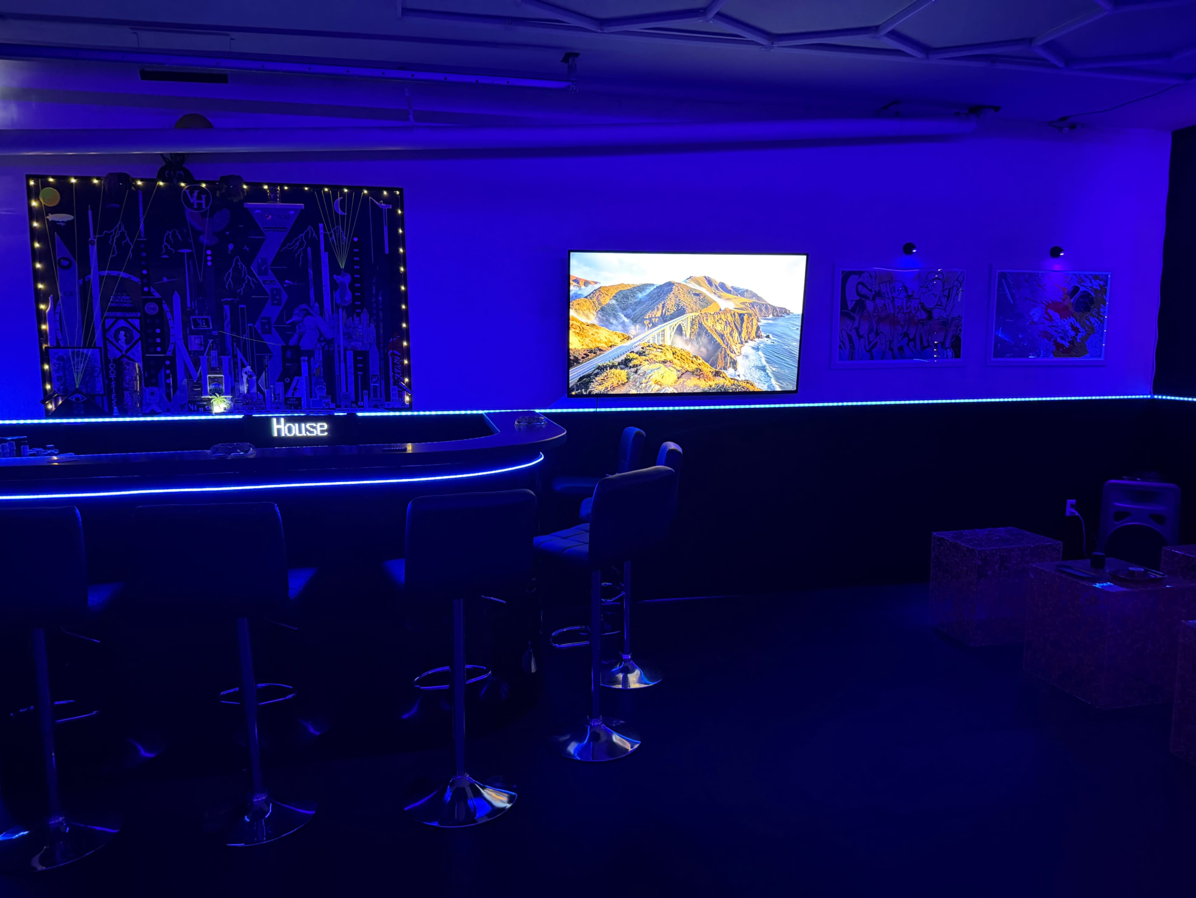 The image shows a dimly lit modern bar area with a blue LED-lit counter, a large flat-screen TV displaying a landscape, and a few art pieces on the walls.