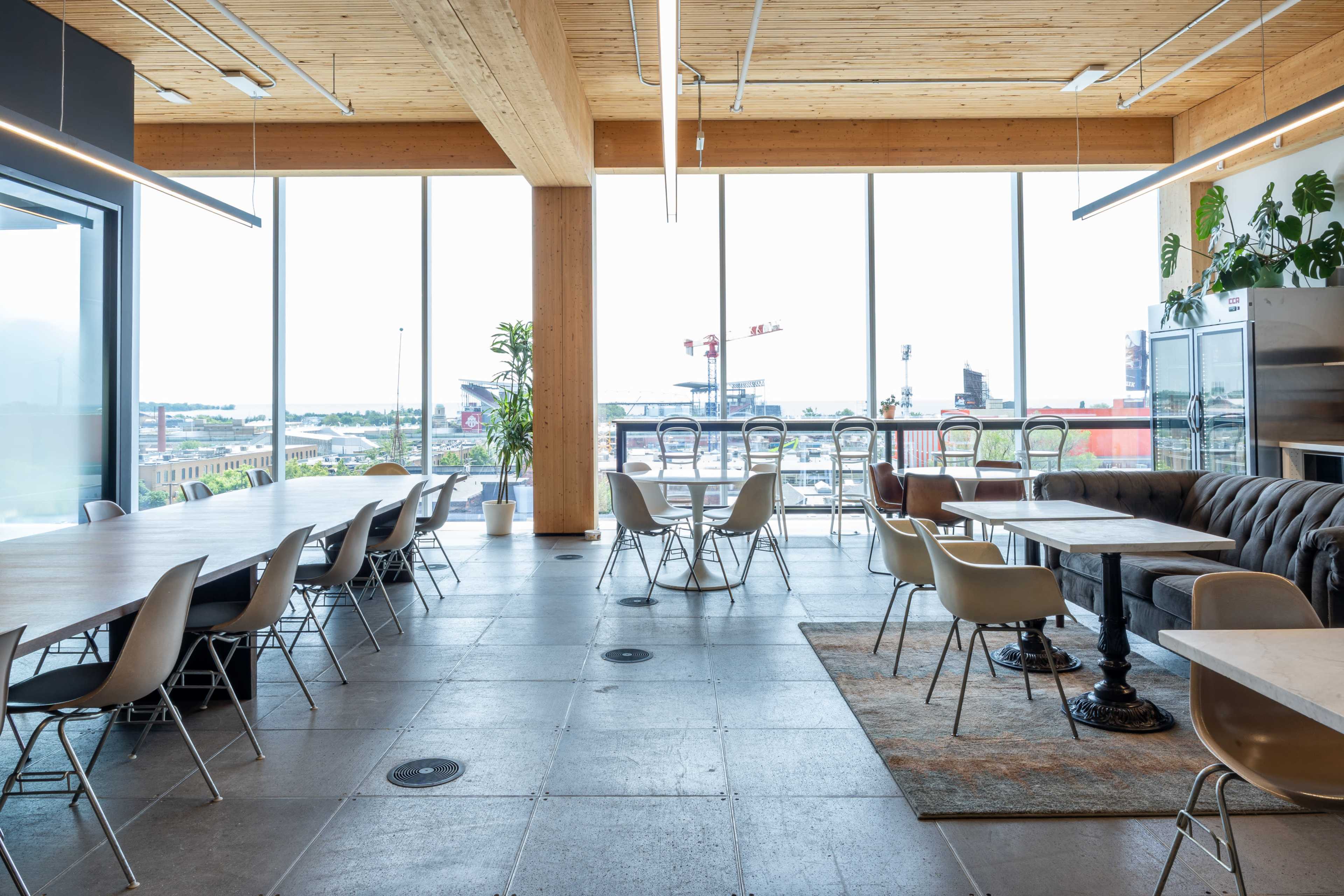The image shows a spacious and modern cafe or meeting area with large windows, wooden beams, and various seating arrangements.
