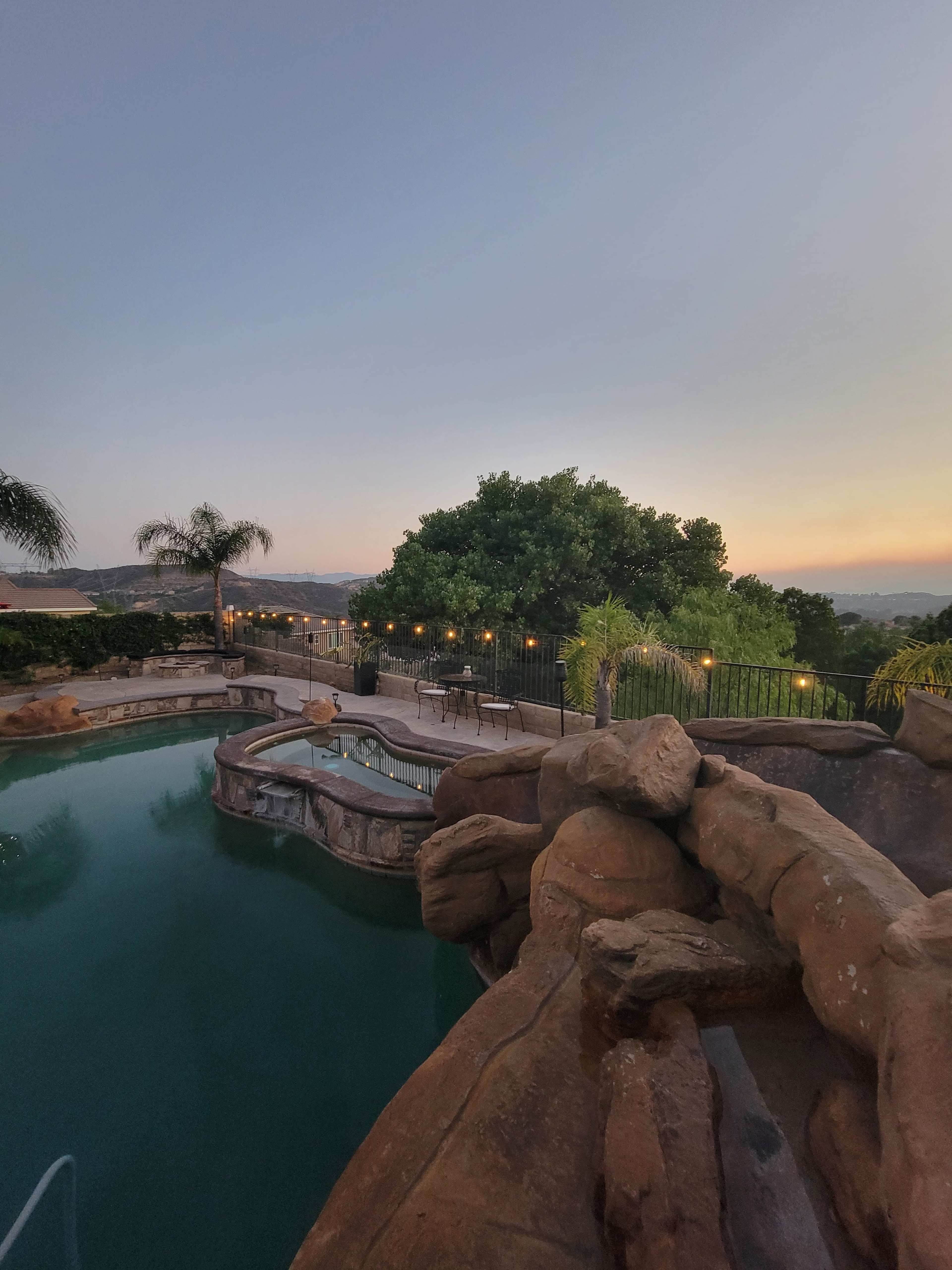 A landscaped backyard features a pool with a rocky slide, surrounded by palm trees and illuminated by string lights at dusk.