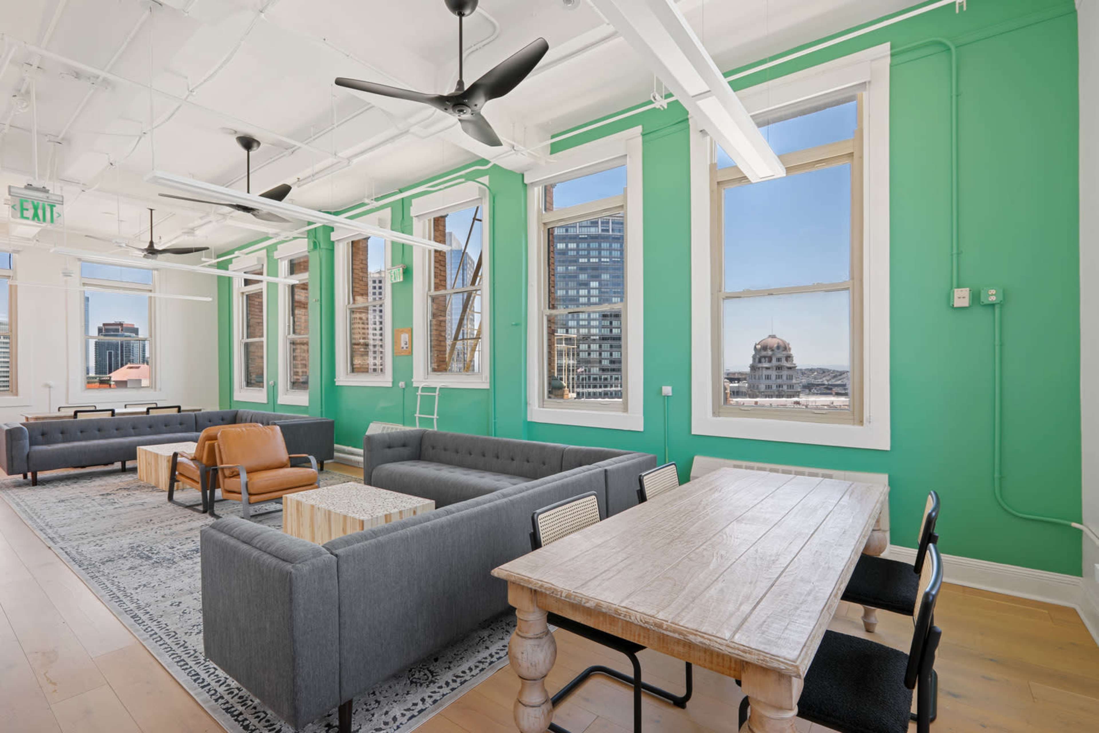 A modern office space with green walls, large windows, and various seating arrangements including sofas and a dining table.