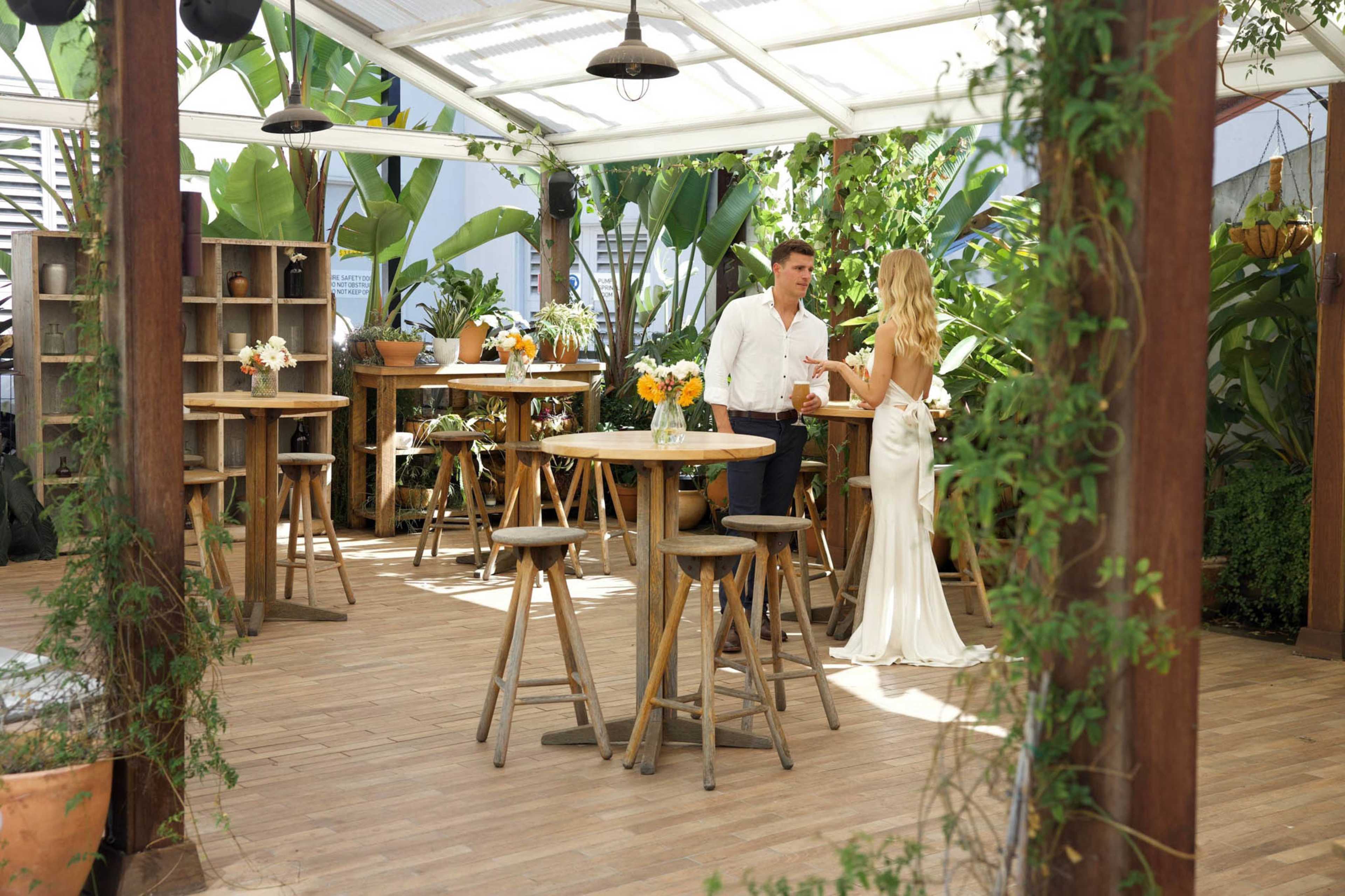 A man and a woman converse in a glass-covered outdoor space filled with plants and wooden furniture.