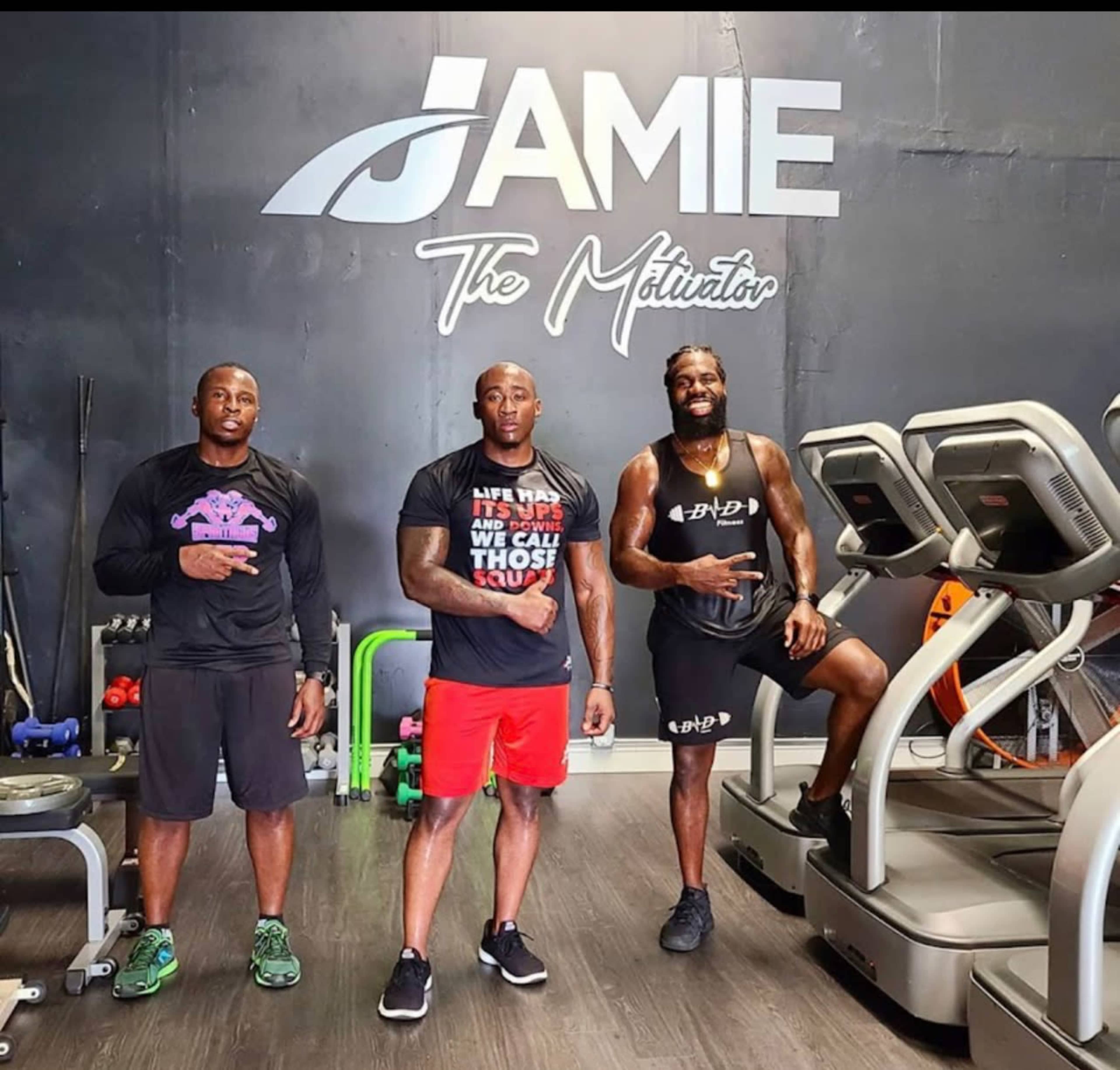 Three men pose in a gym with fitness equipment and a wall sign that reads "JAMIE The Motivator."