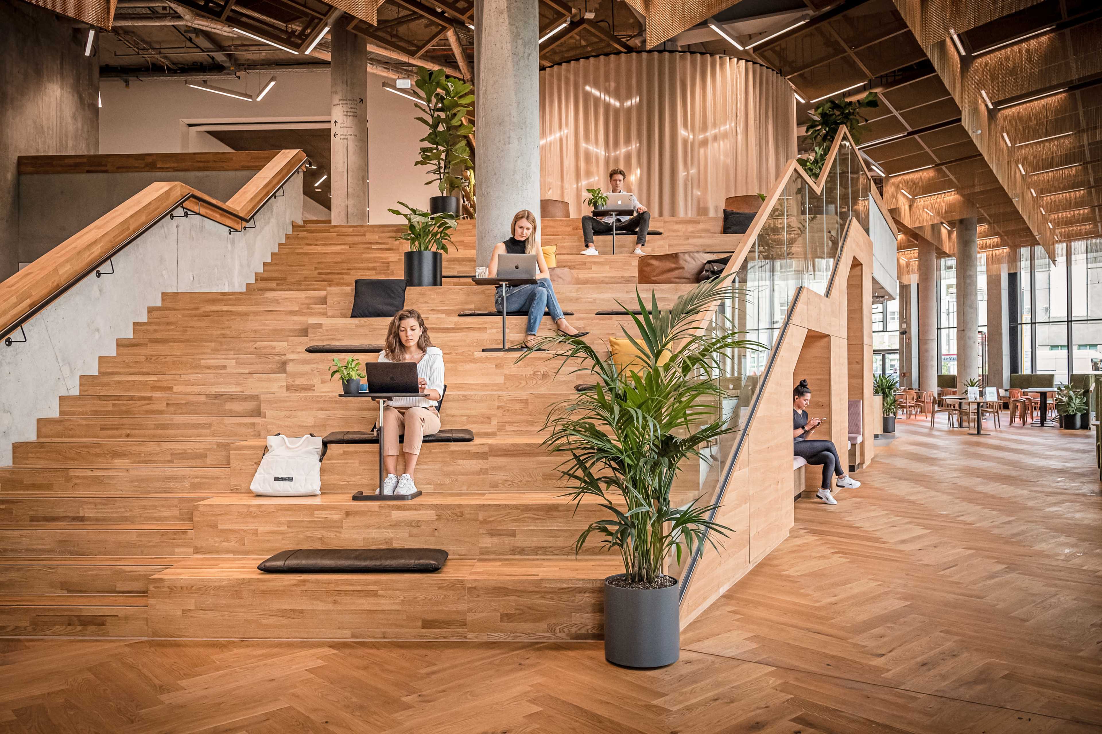 A group of people are working on laptops while seated on wooden steps in a modern, open office space surrounded by plants.