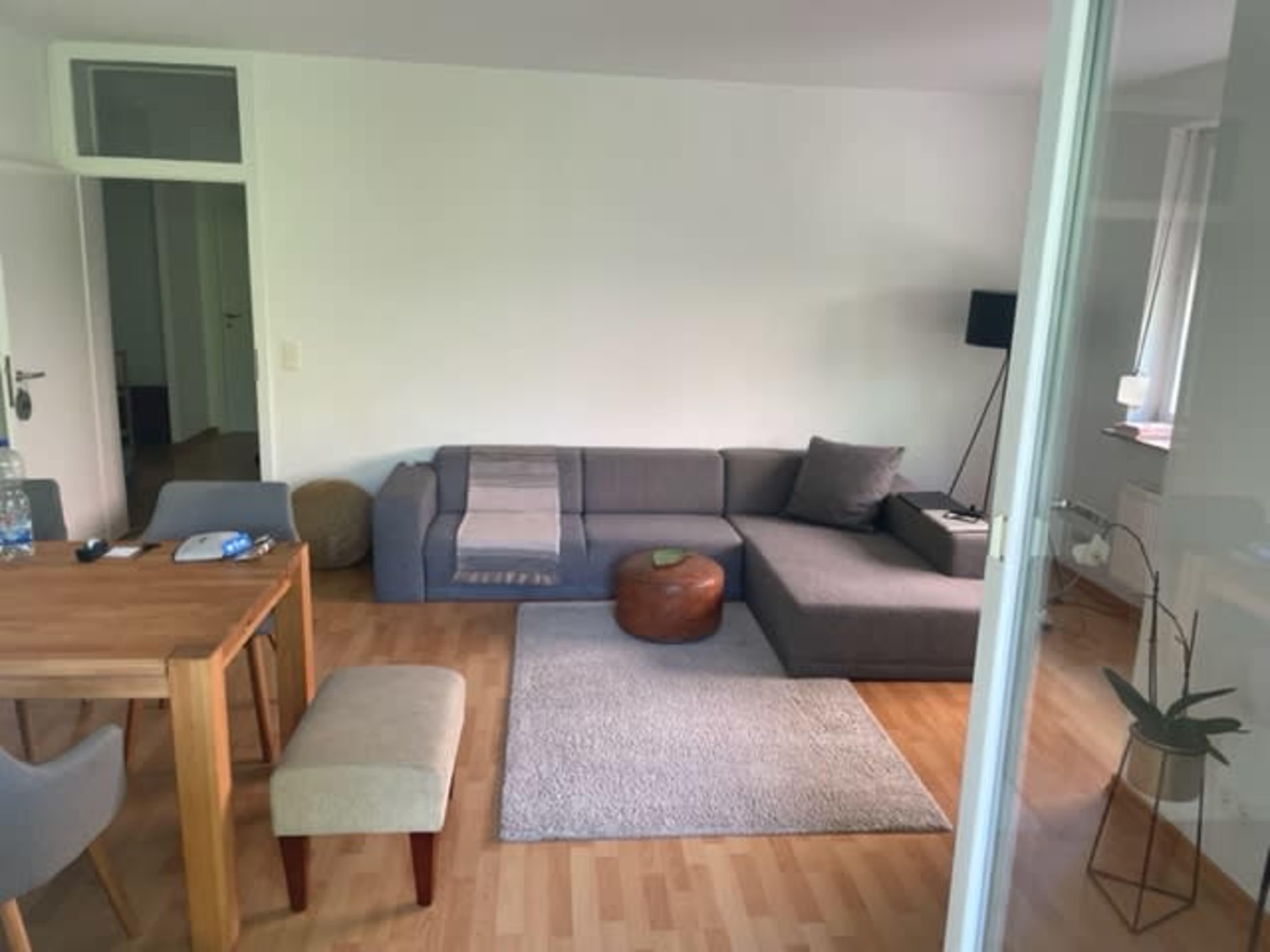 A modern living room with a gray sectional sofa, a wooden dining table, a small stool, and a rug on a hardwood floor.