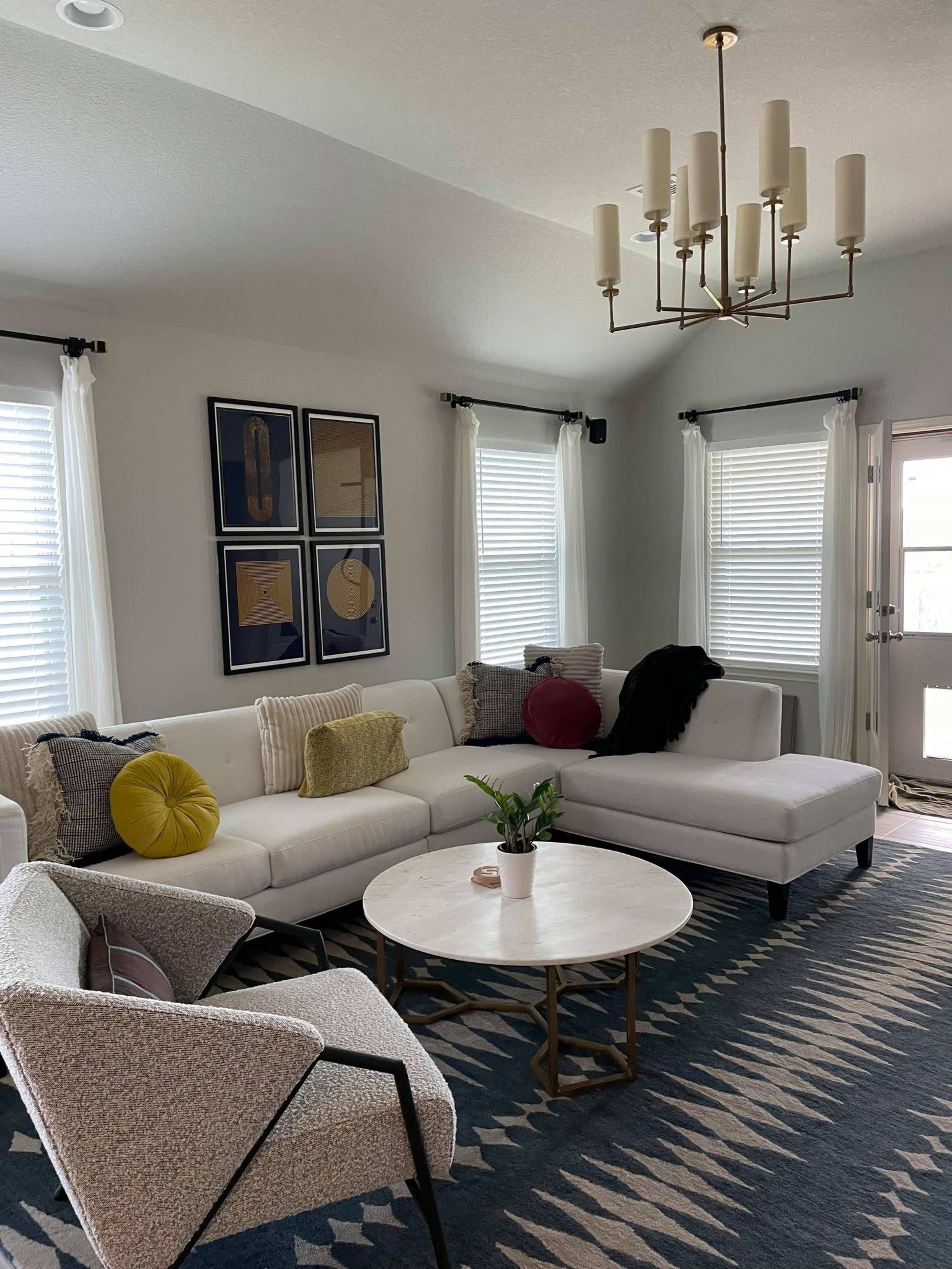 A modern living room features a white sectional sofa, a round marble coffee table, and a patterned rug, with artwork on the walls and large windows letting in natural light.