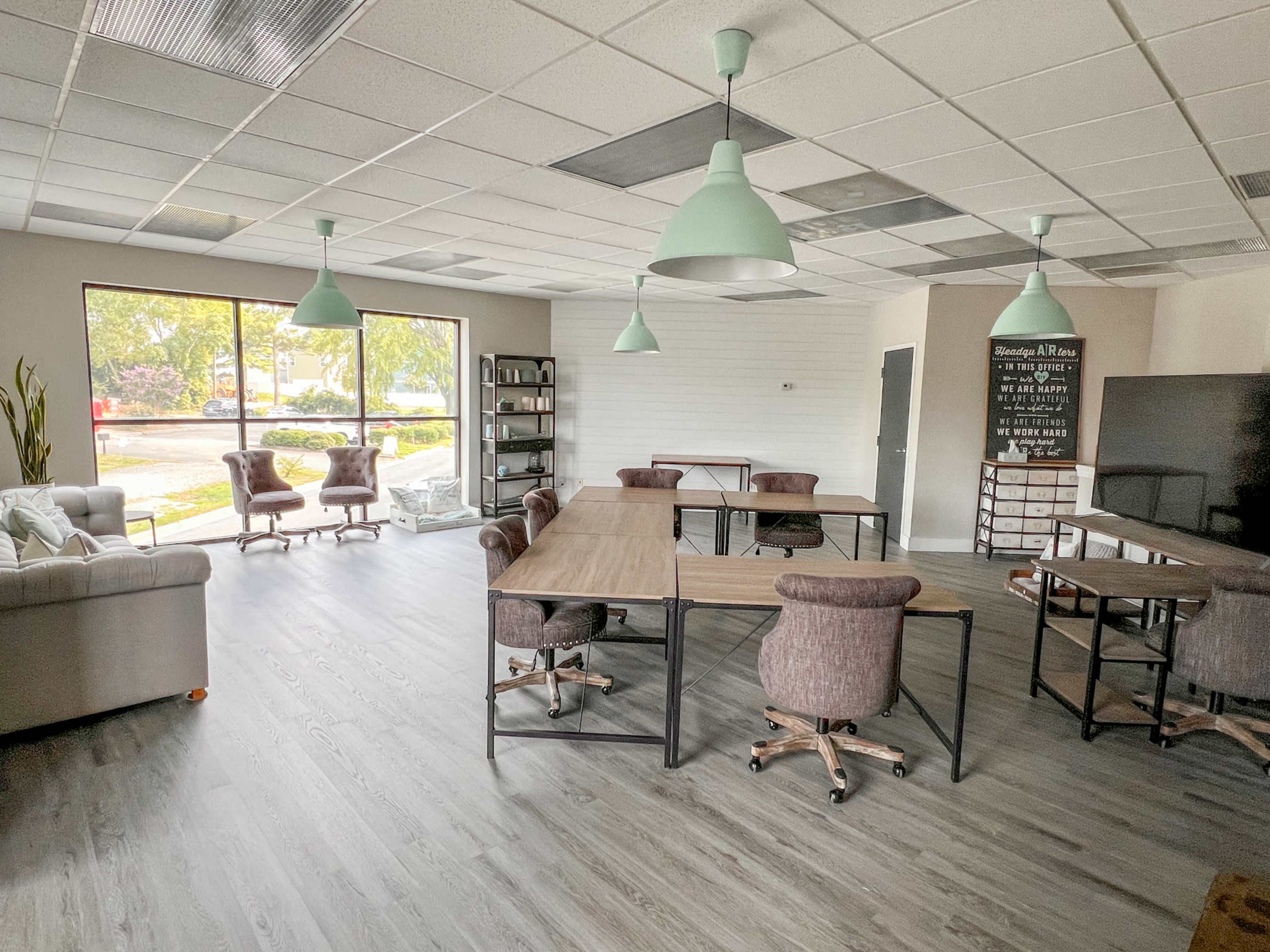The image shows a modern office interior with several tables and chairs, a bookshelf, and large windows allowing natural light to enter.