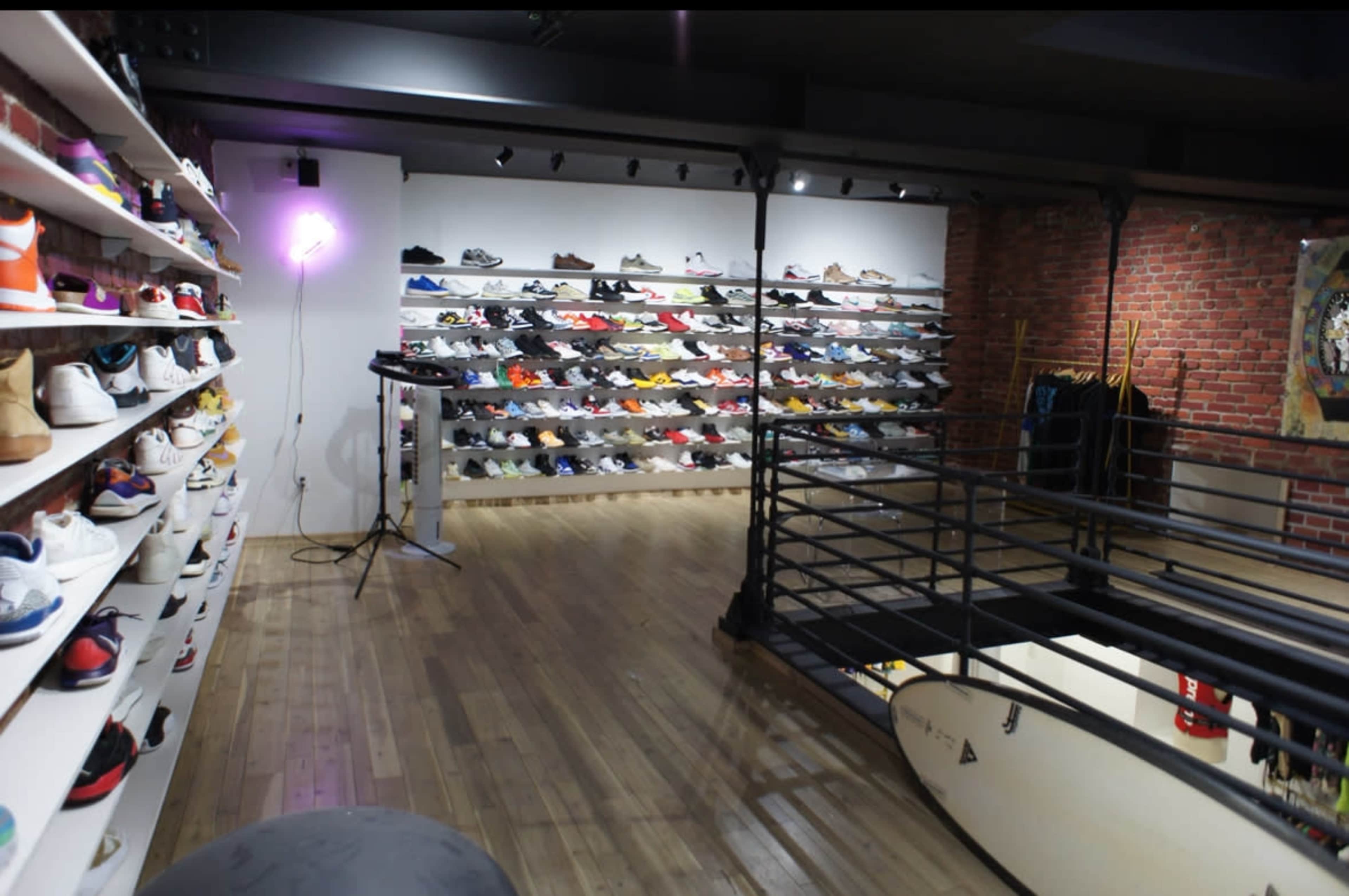 A shoe boutique displays a wide variety of sneakers on shelves against a brick wall, with a surfboard and equipment in the foreground.