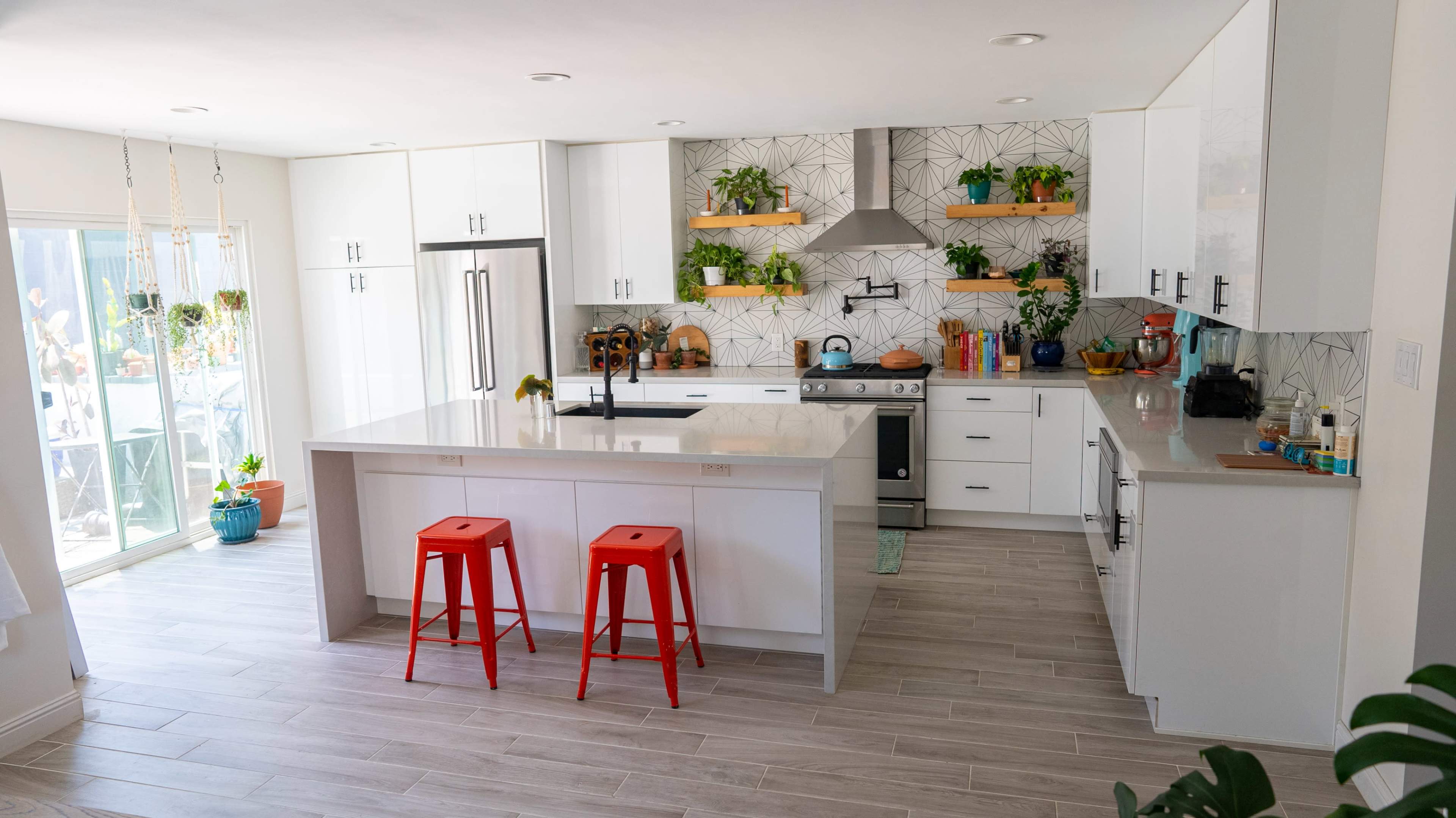 The image shows a modern kitchen with white cabinets, a central island with red stools, and plants arranged on shelves and countertops.