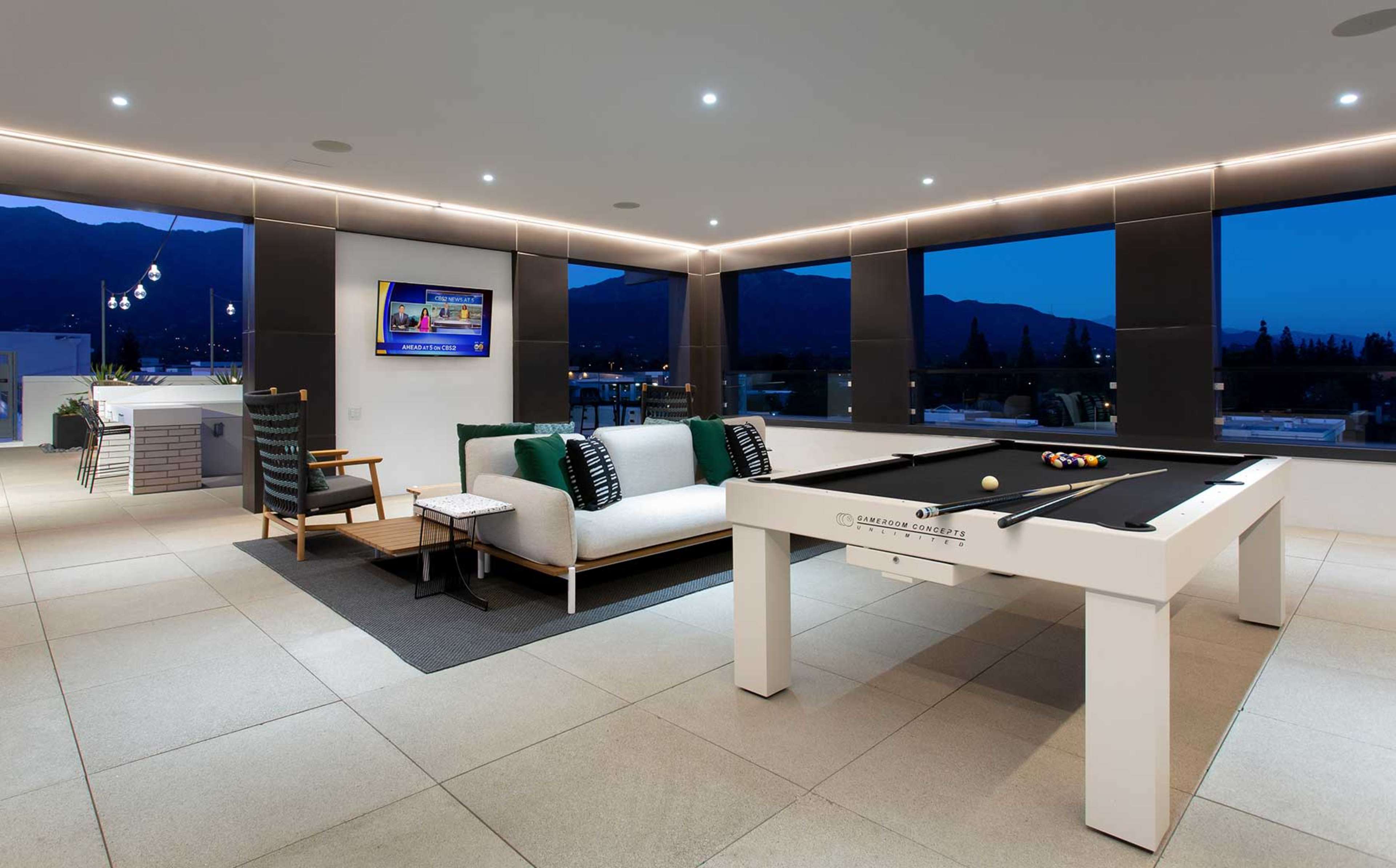 A modern lounge area featuring a billiard table, a sectional sofa, and large windows with a view of mountains at dusk.