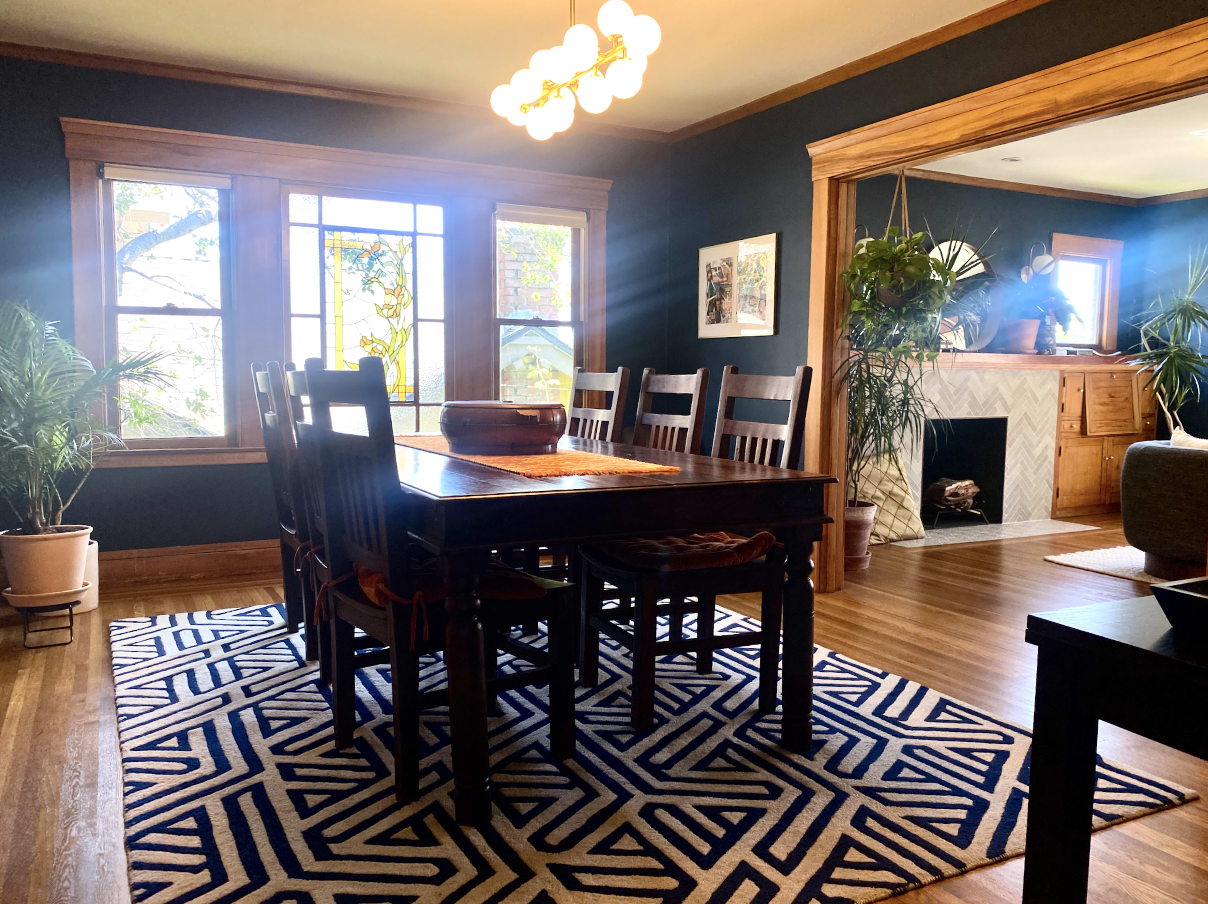 The image shows a dining room featuring a large wooden table surrounded by chairs, with a patterned rug on the floor and large windows allowing natural light to enter.
