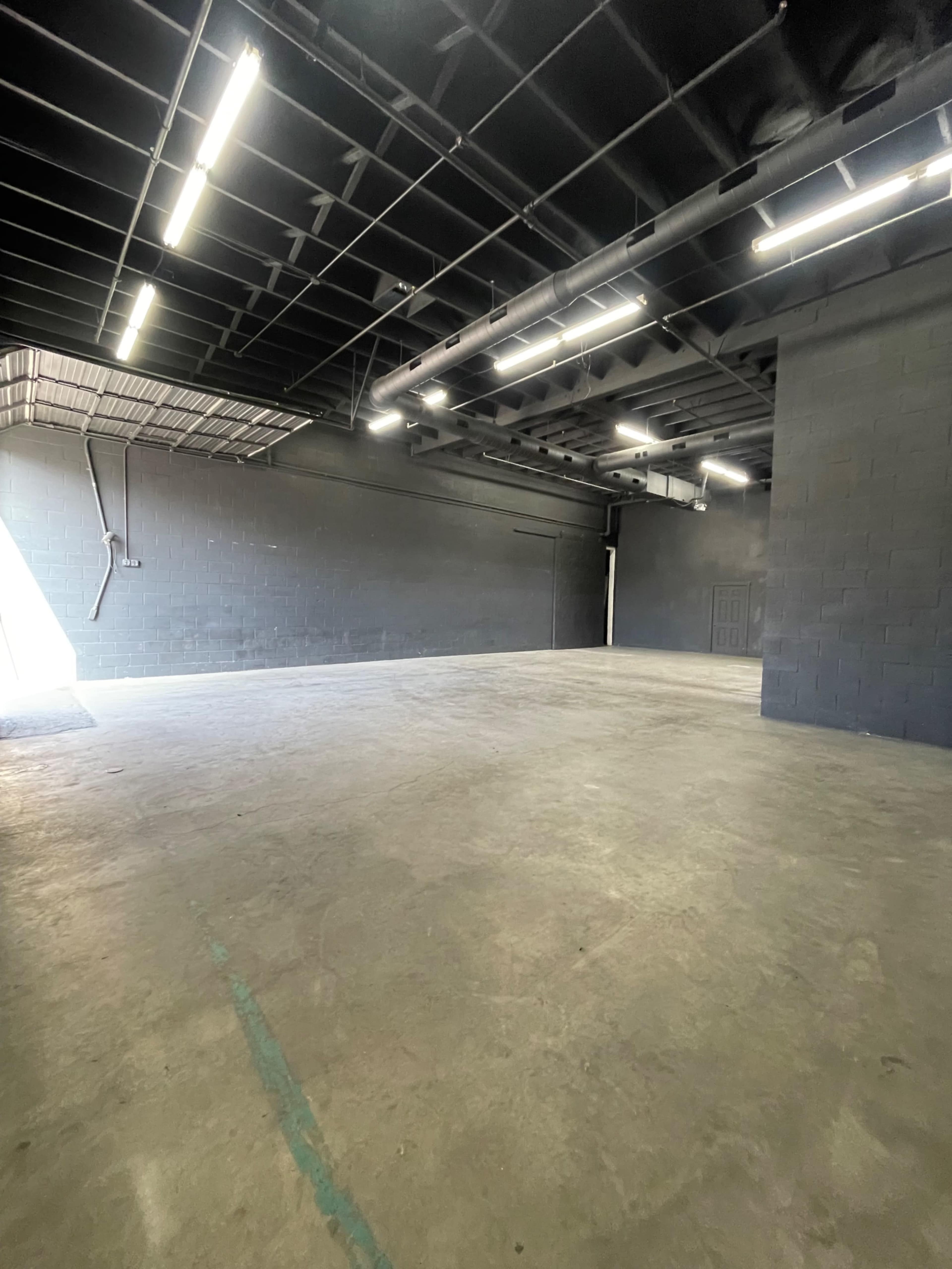 The image shows a large, empty industrial space with concrete floors and black walls, illuminated by fluorescent lighting.