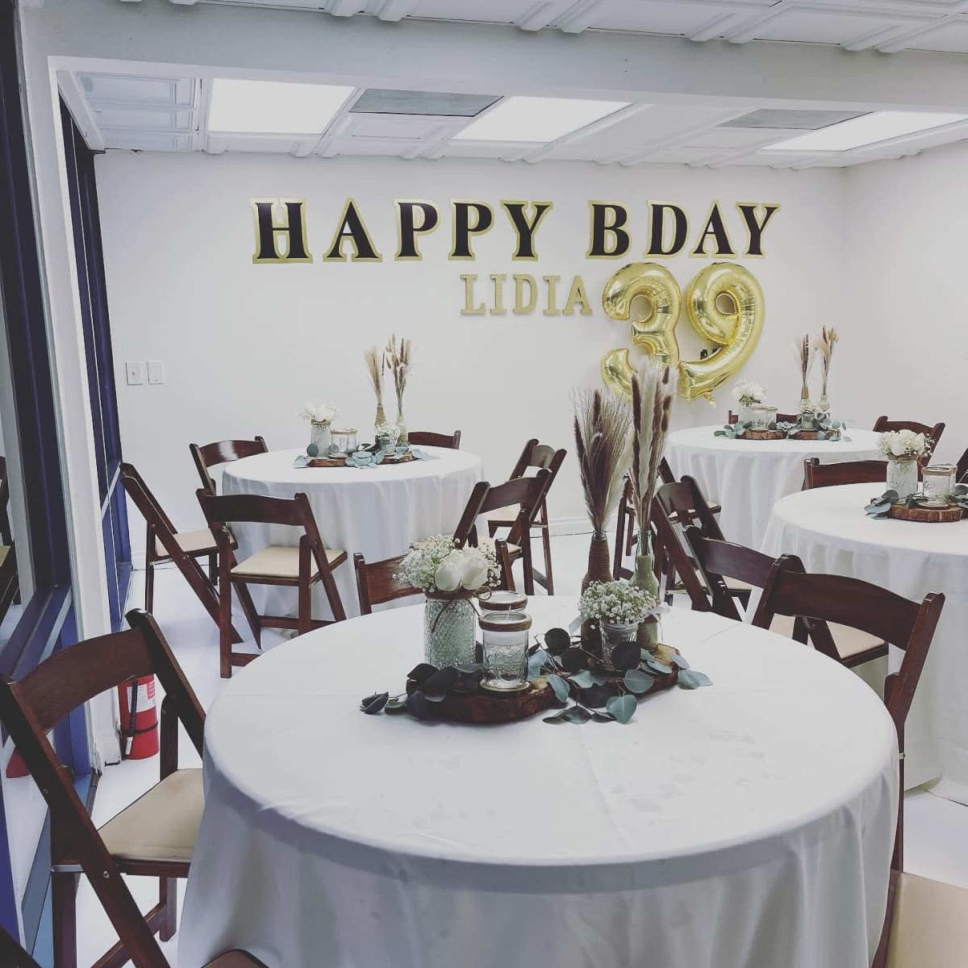 The image shows a decorated room with round tables covered in white tablecloths, featuring floral centerpieces, and a wall displaying a "HAPPY BDAY LIDIA 39" banner and gold balloons.