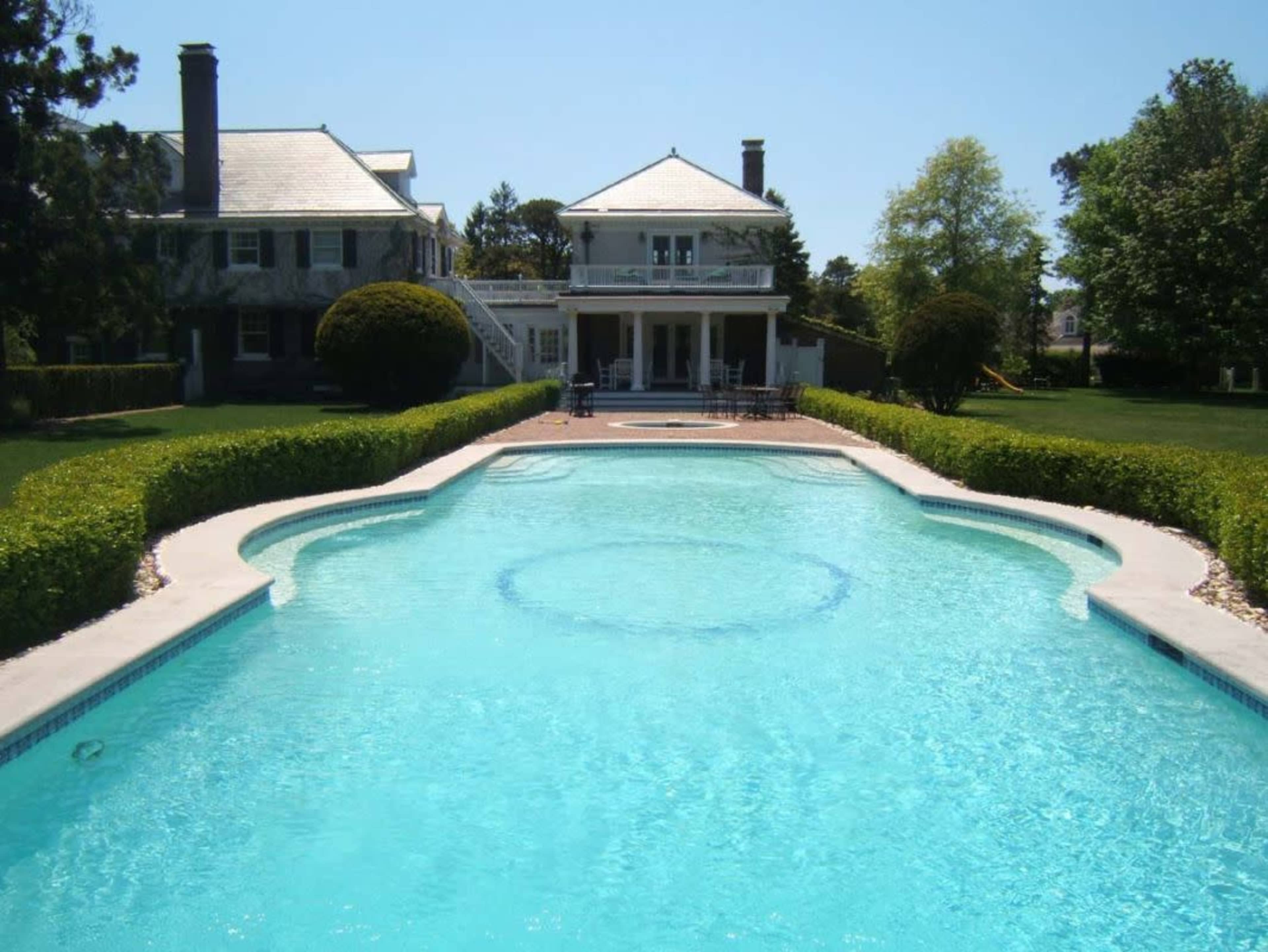 A clear blue swimming pool is situated in front of a large house, surrounded by well-manicured lawns and hedges.