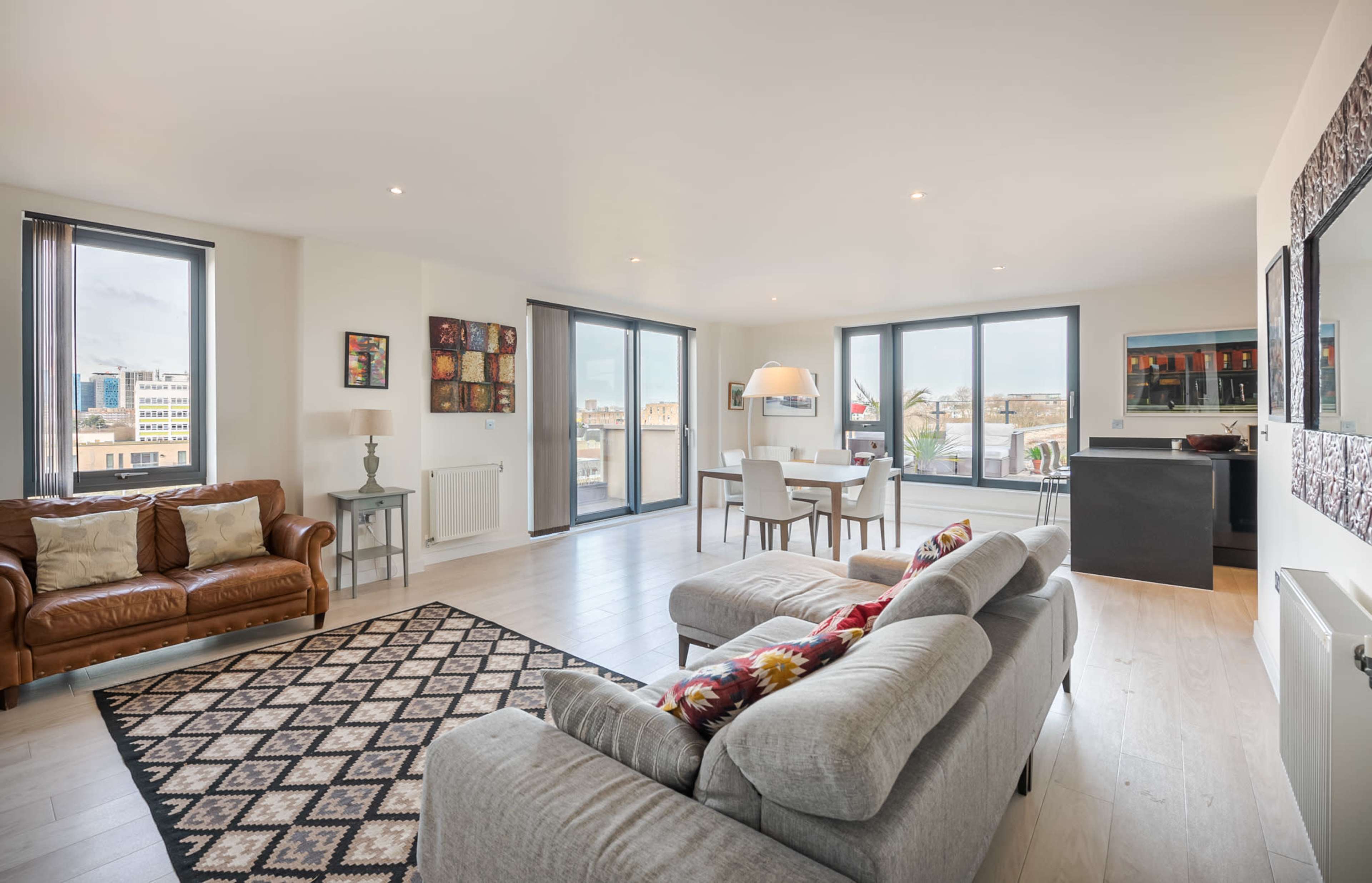 The image shows a modern, open-concept living area with a leather couch, a patterned rug, and large windows allowing natural light to fill the space.