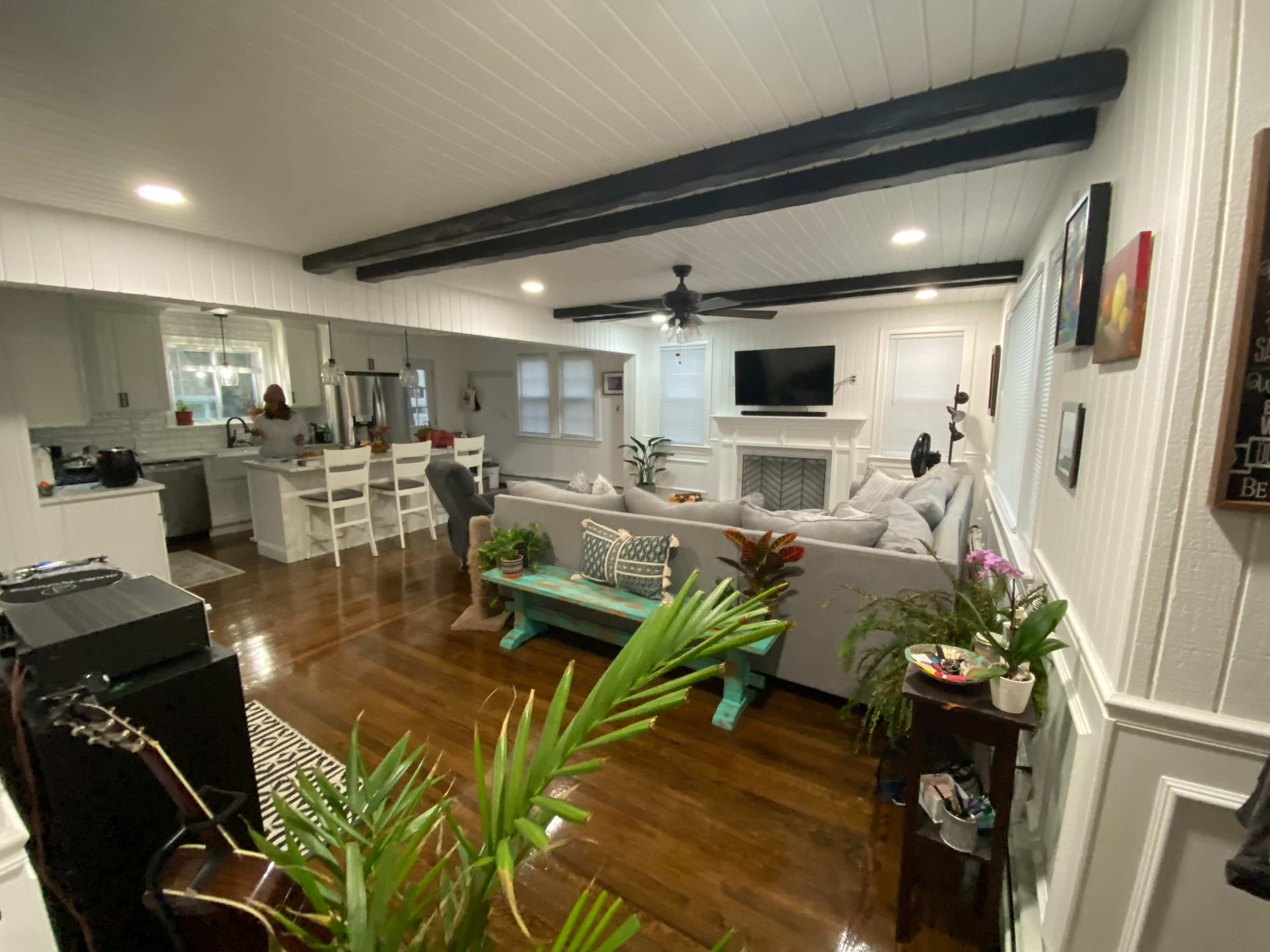 A cozy living room with a gray sectional sofa, wooden beams on the ceiling, and a kitchen area visible in the background.