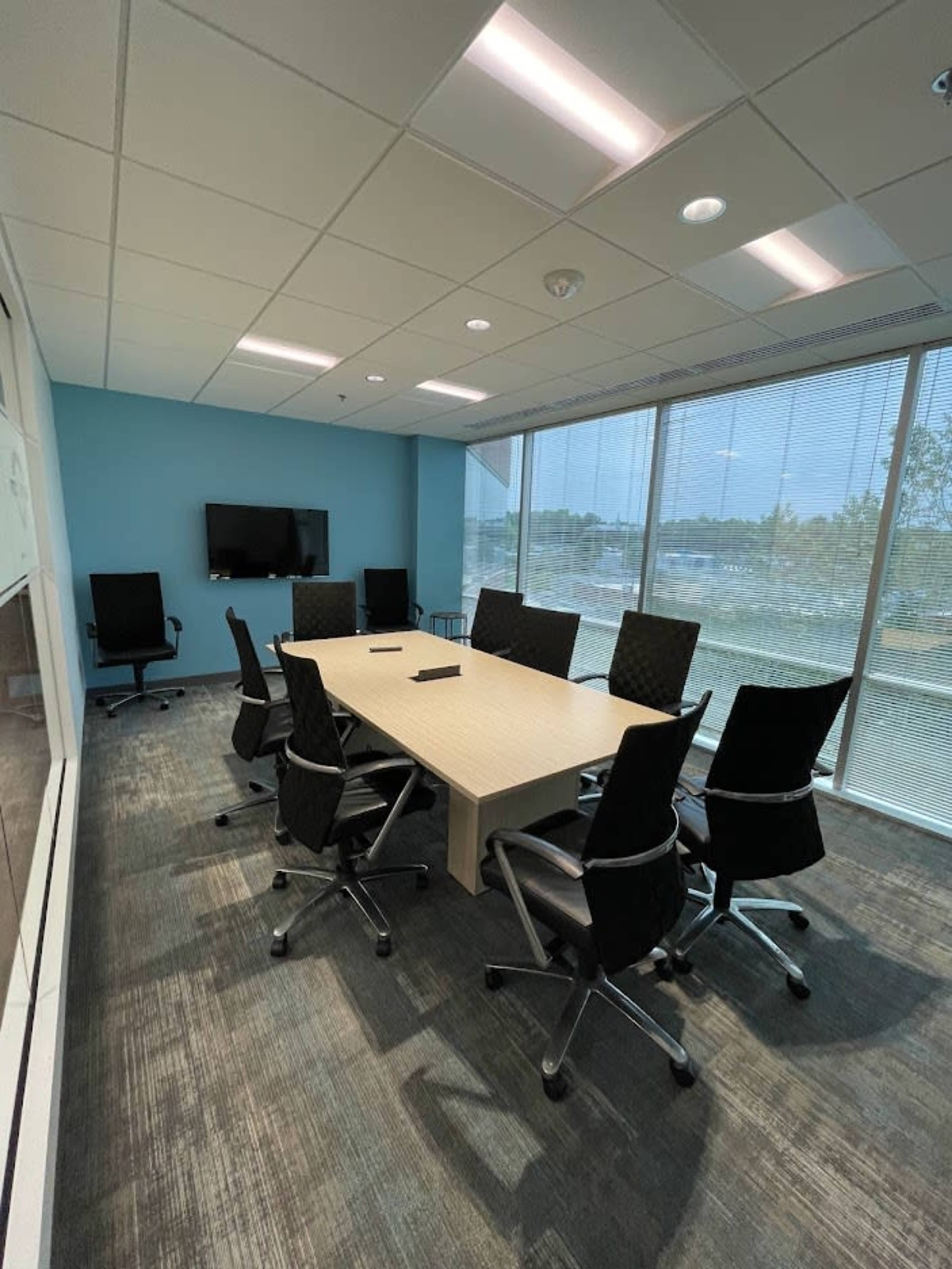 A conference room features a large table surrounded by ergonomic chairs, with a television mounted on the wall and windows letting in natural light.