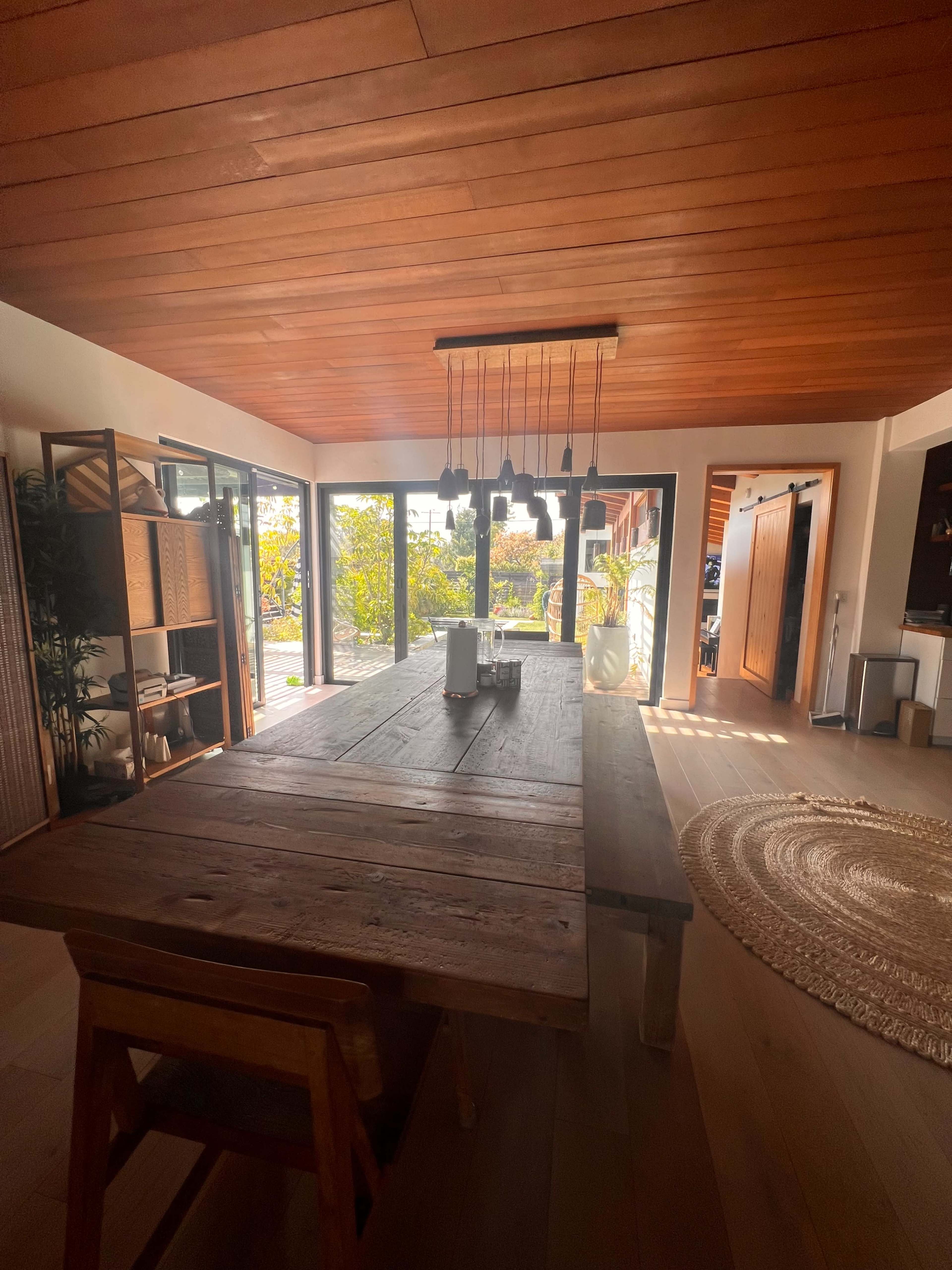 A large wooden dining table sits in a brightly lit room with expansive glass doors leading to an outdoor space.