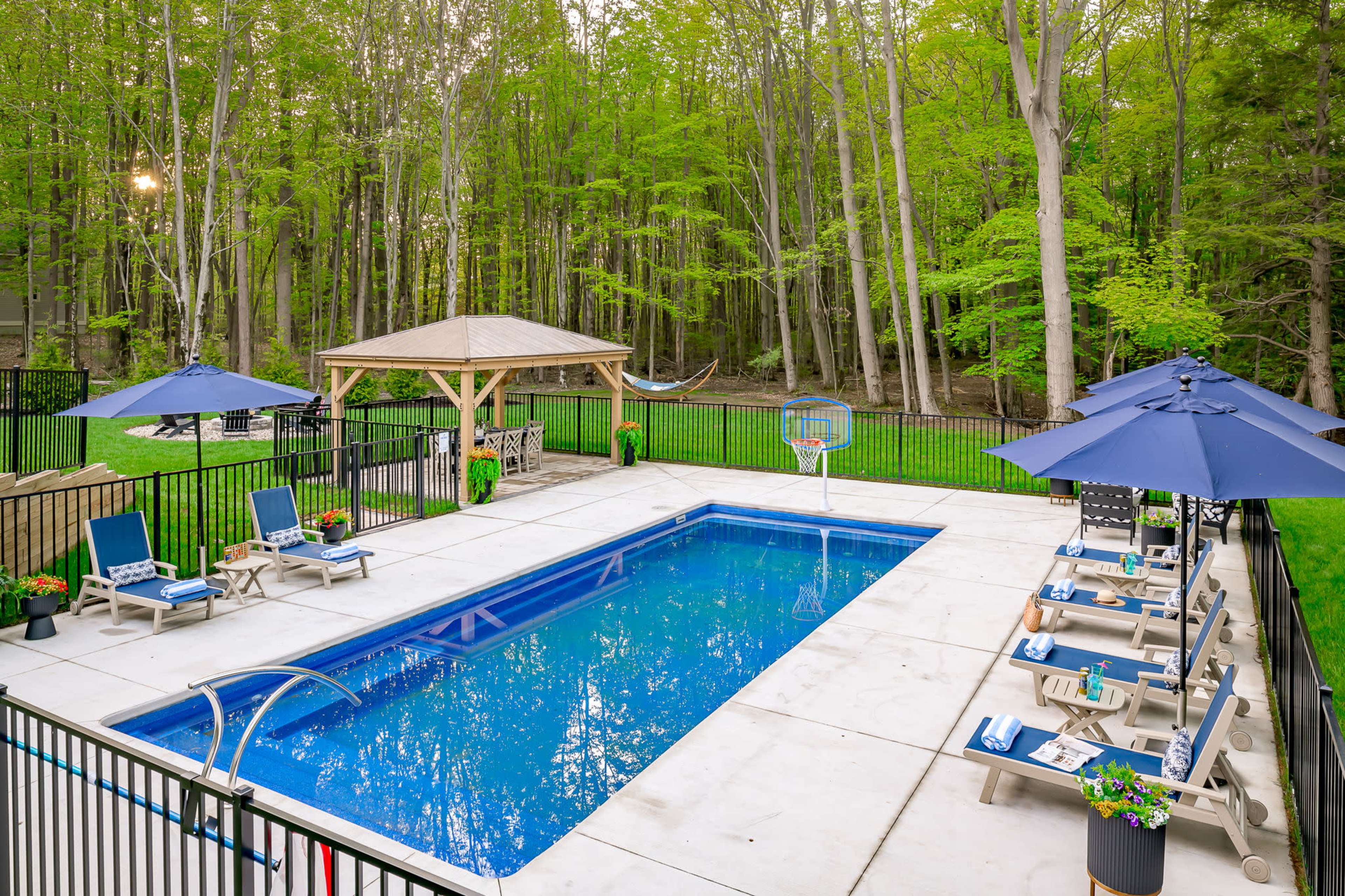 A rectangular swimming pool surrounded by lounge chairs and umbrellas, set in a green, wooded backyard with a gazebo and a hammock.