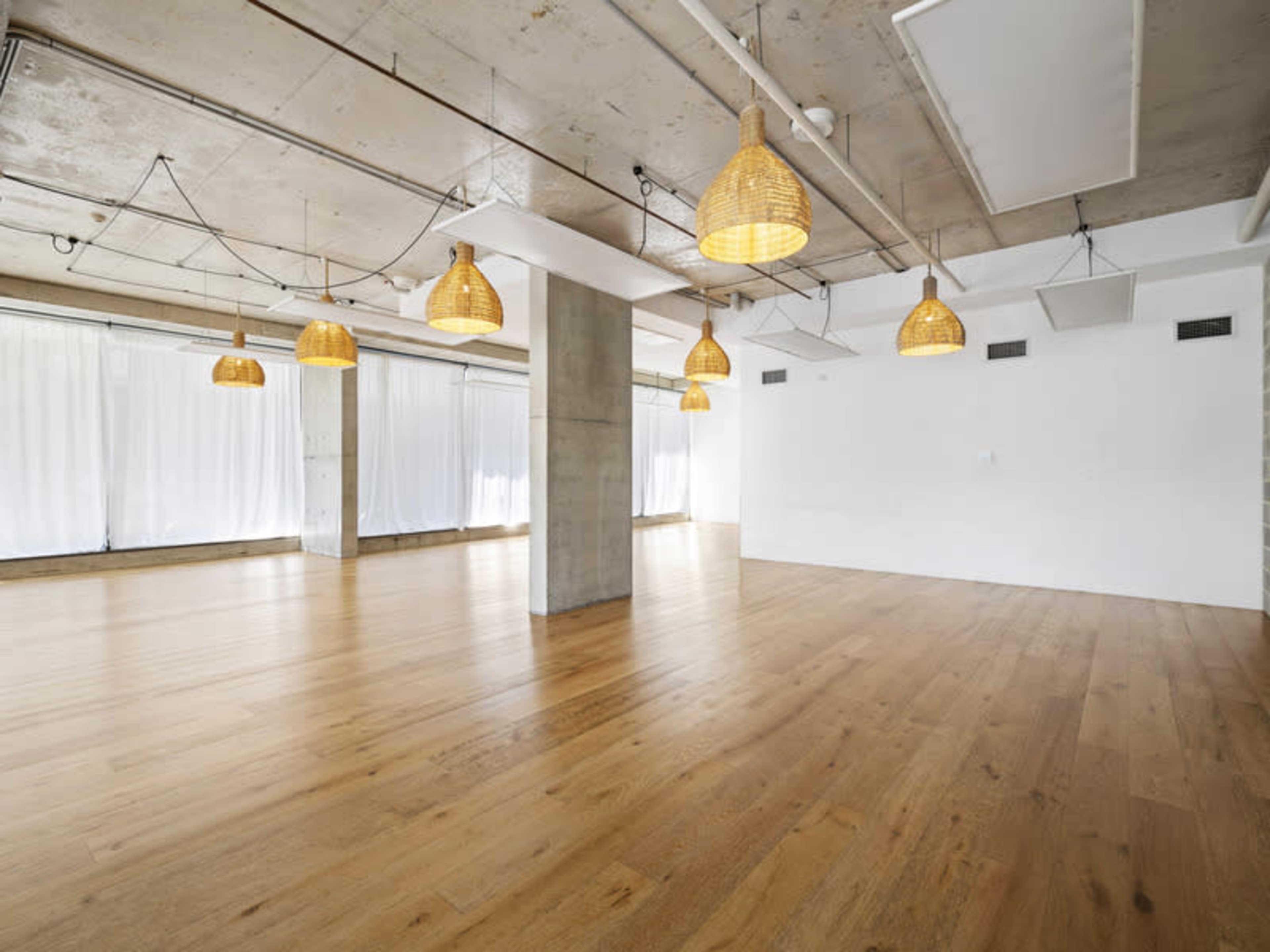 The image shows a spacious, empty room featuring wooden flooring, large windows with white curtains, and pendant lights hanging from the ceiling.