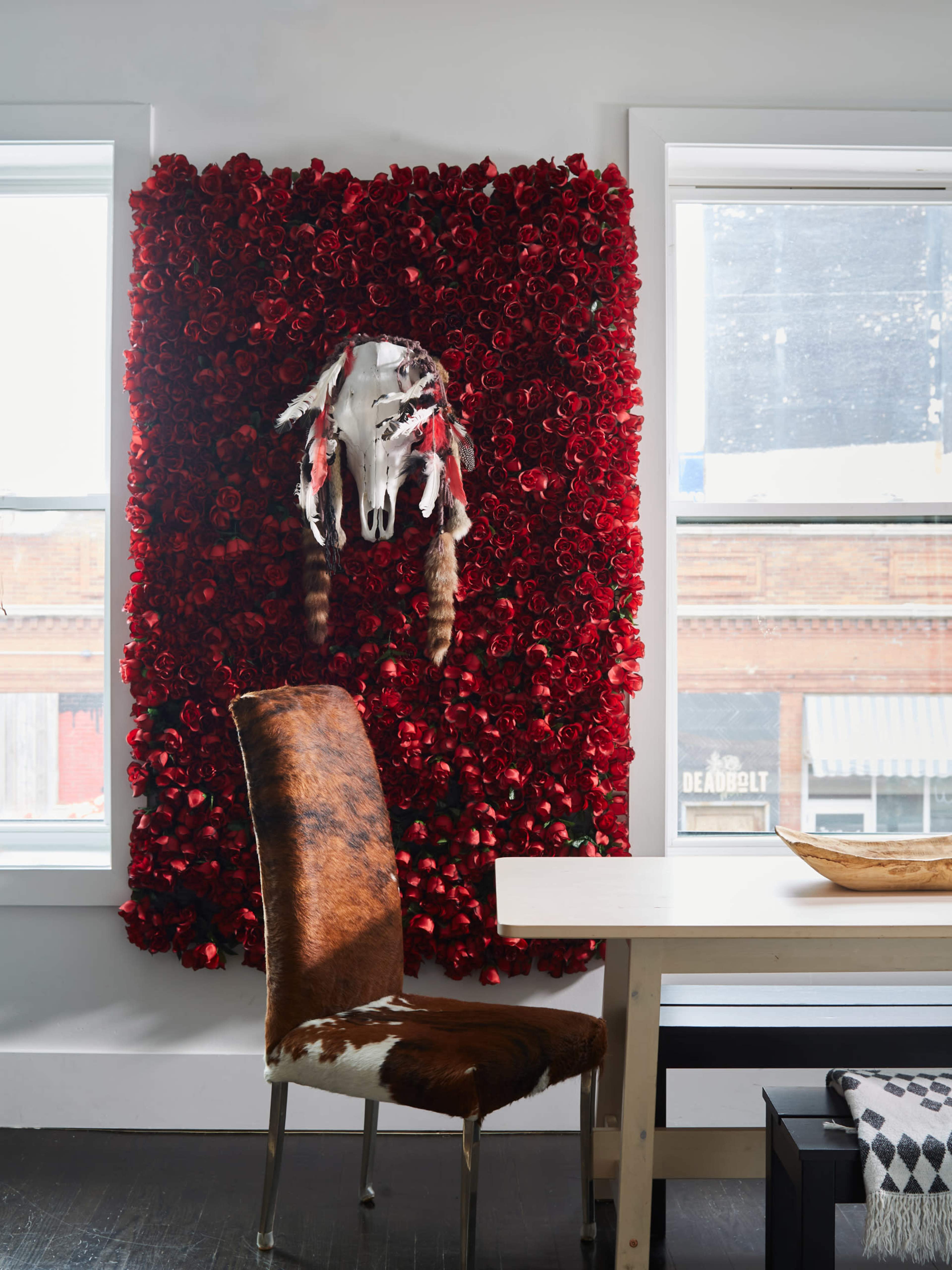 A wall covered in red roses features a decorative cow skull with feathers above a wooden table and a chair with a cowhide pattern.