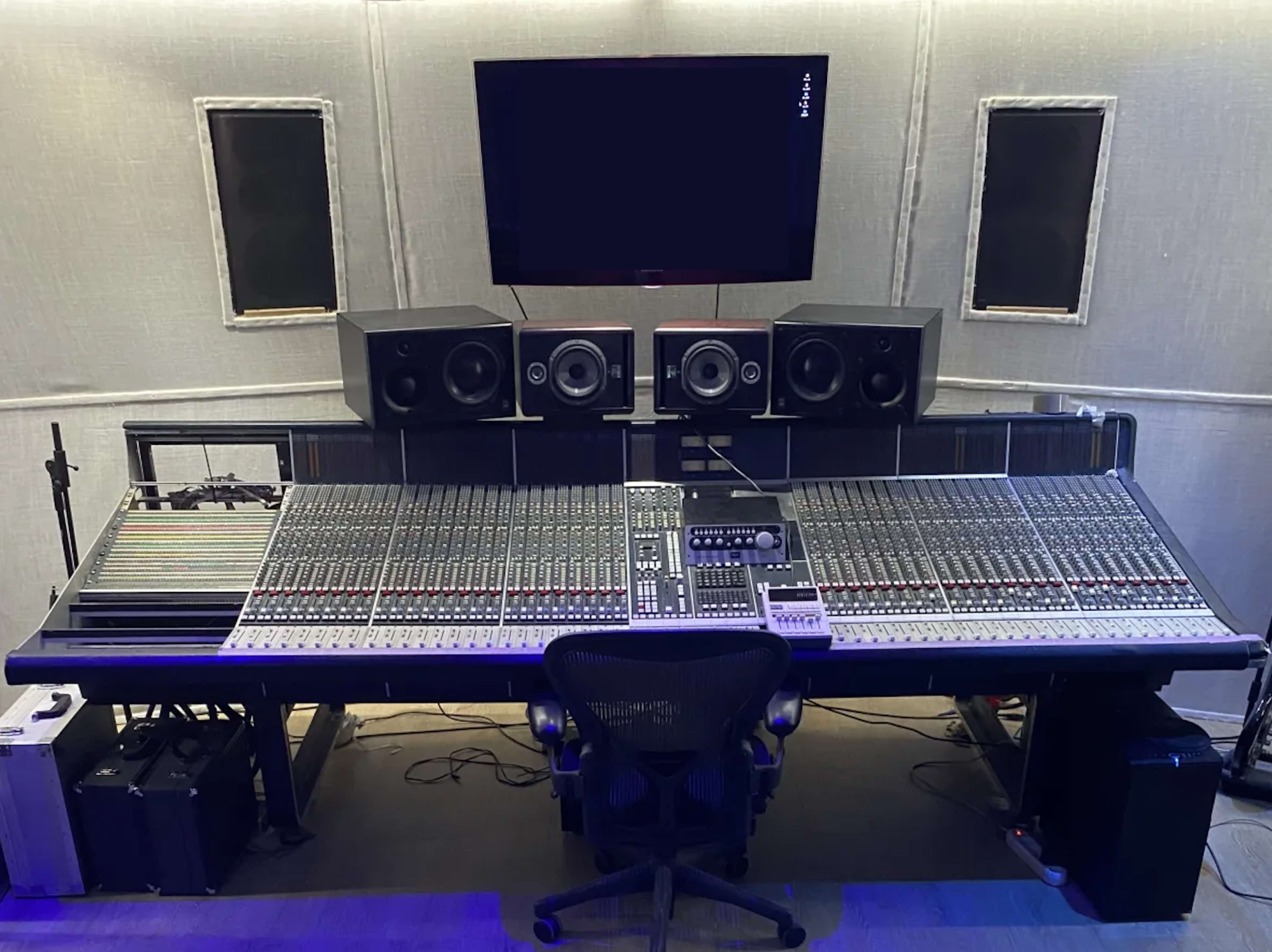 A recording studio with a large mixing console, two studio monitors, and a wall-mounted screen.