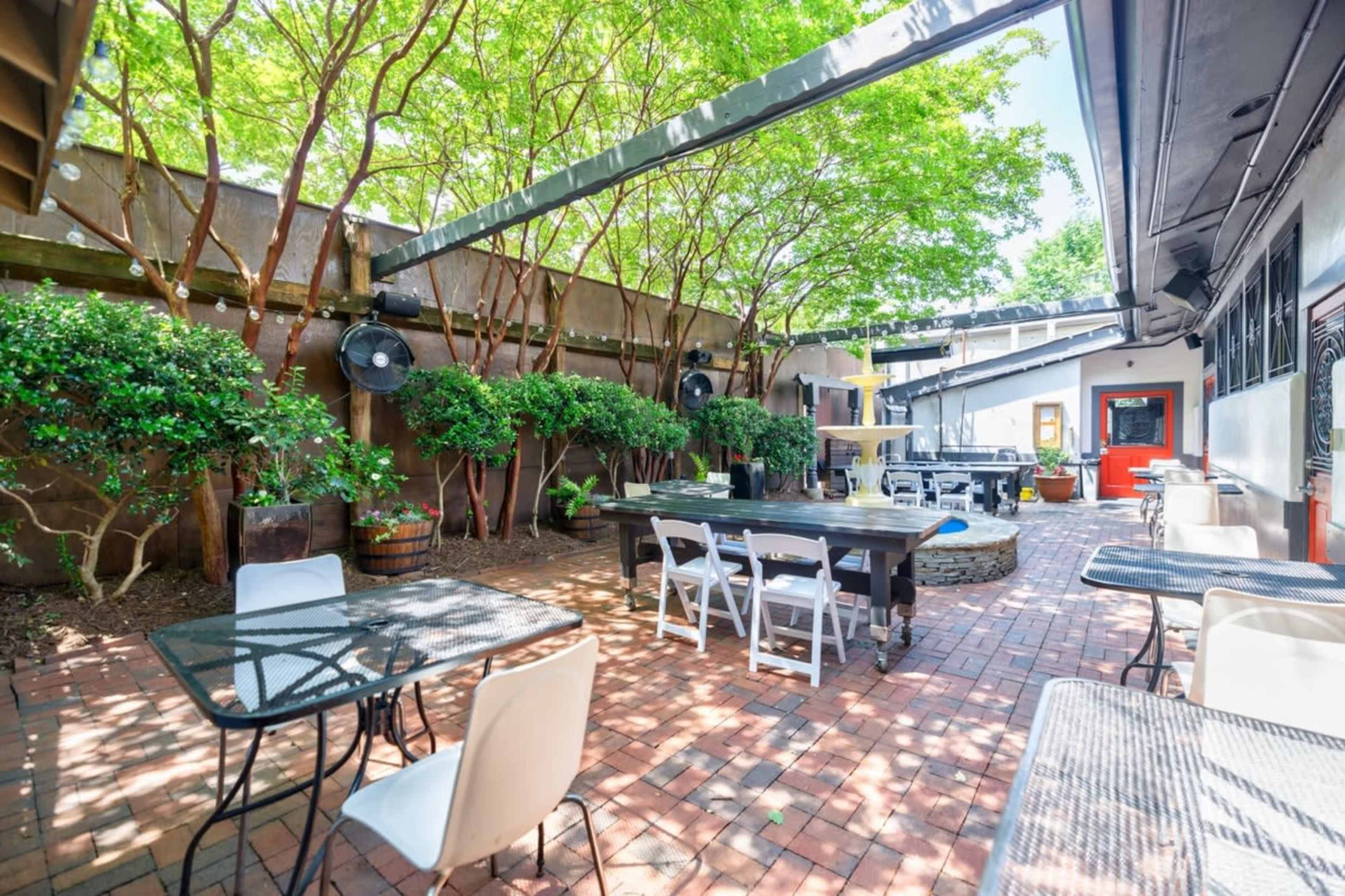 The image shows an outdoor patio area featuring brick flooring, tables, and greenery, with a fountain in the center.
