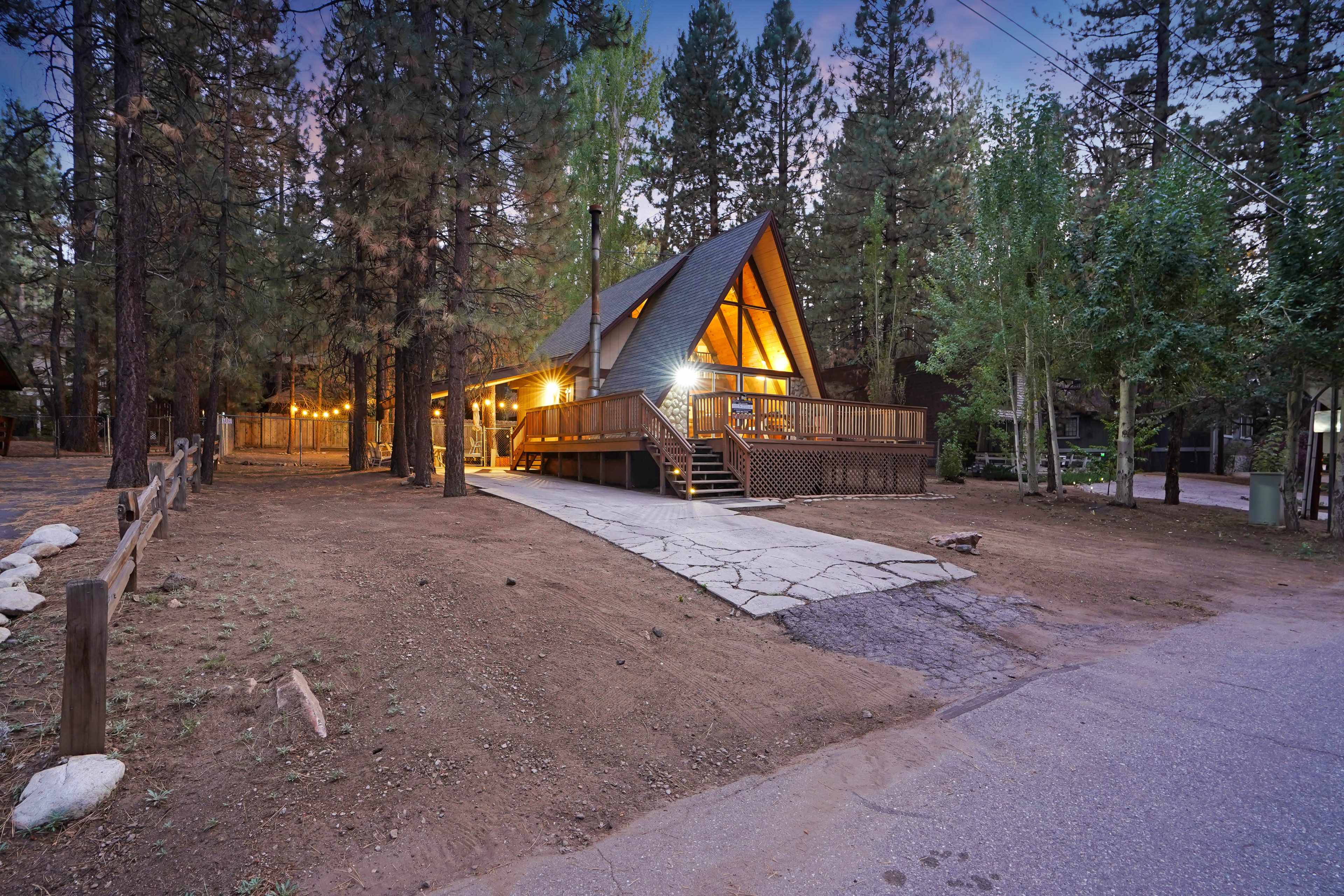 A wooden A-frame cabin with a deck is illuminated at dusk, surrounded by tall pine trees and gravel pathways.