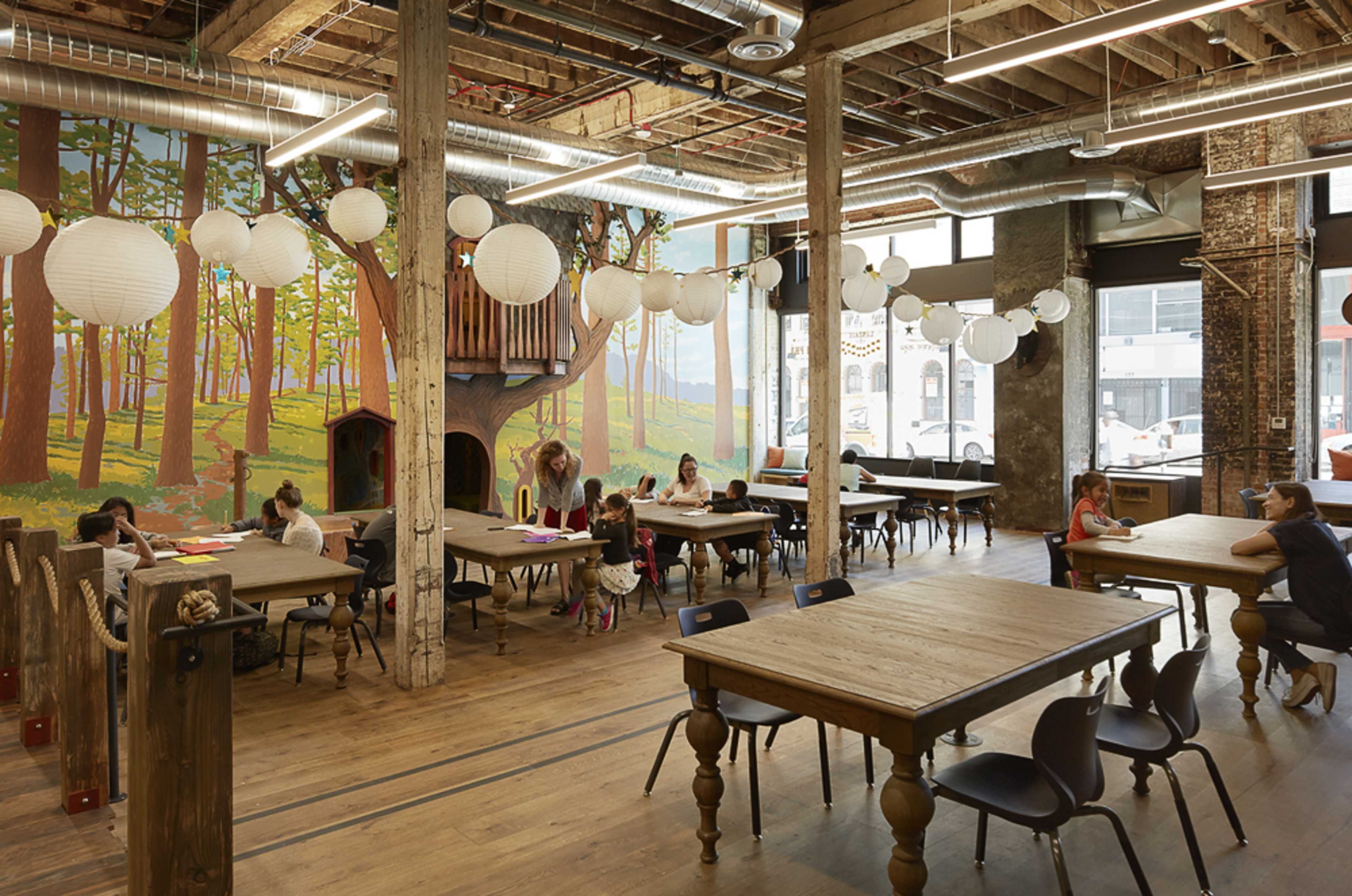 A spacious, well-lit interior of a creative workspace with large tables, wooden flooring, and a mural of trees on the far wall.