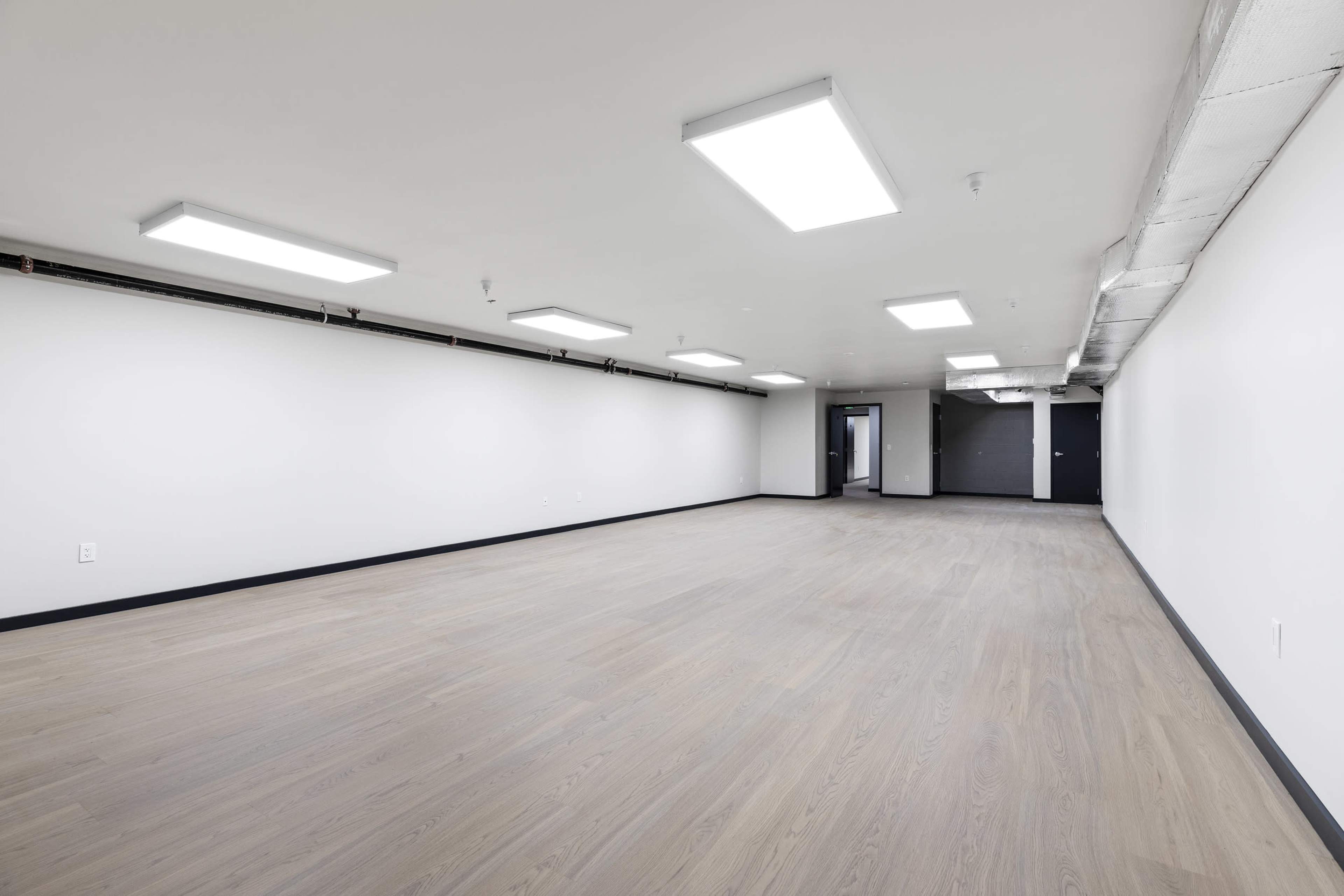 The image shows a spacious, empty room with wooden flooring and fluorescent ceiling lights.