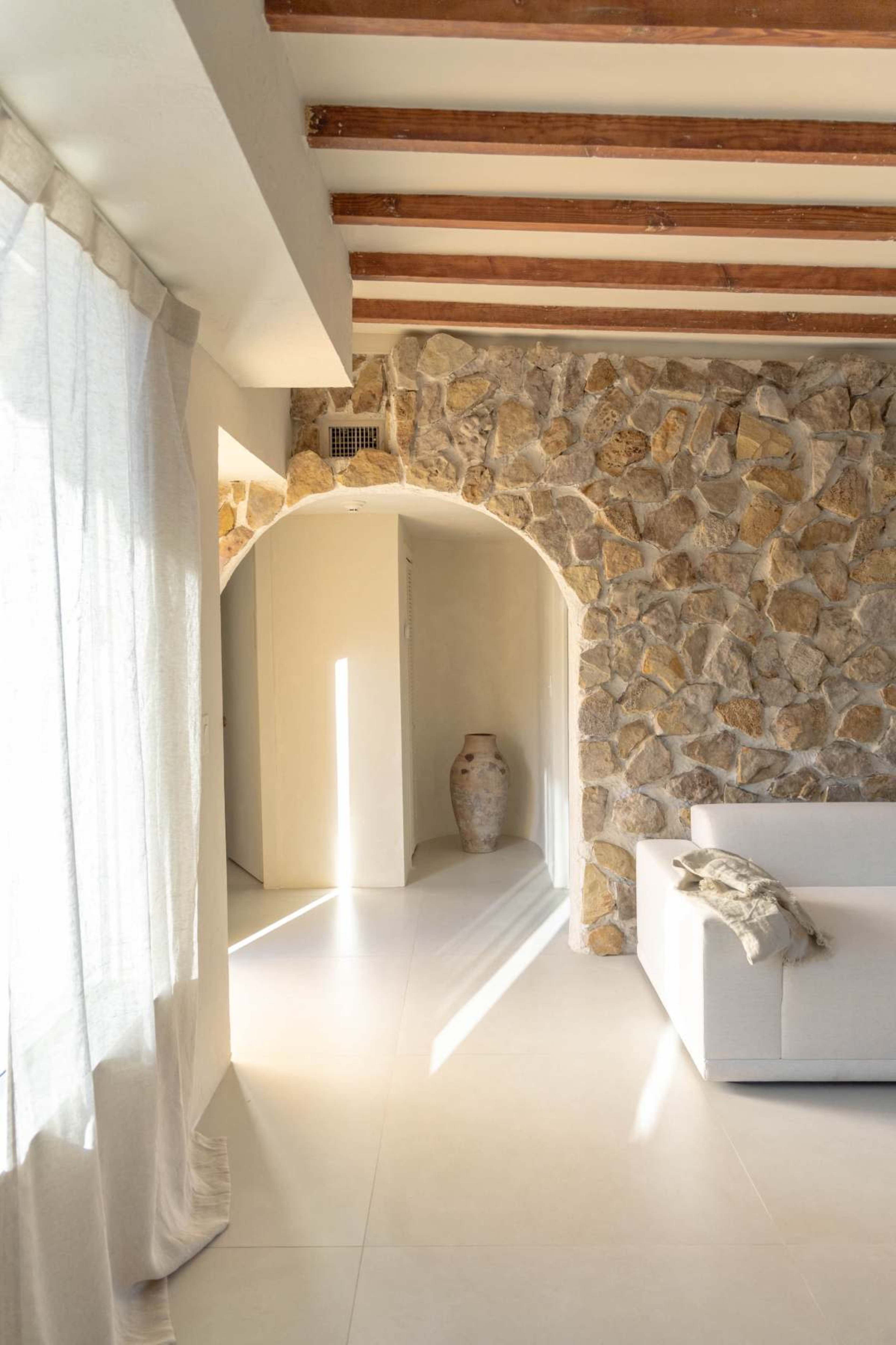 The image shows a bright and airy living space with stone walls, wooden ceiling beams, and a modern white sofa.