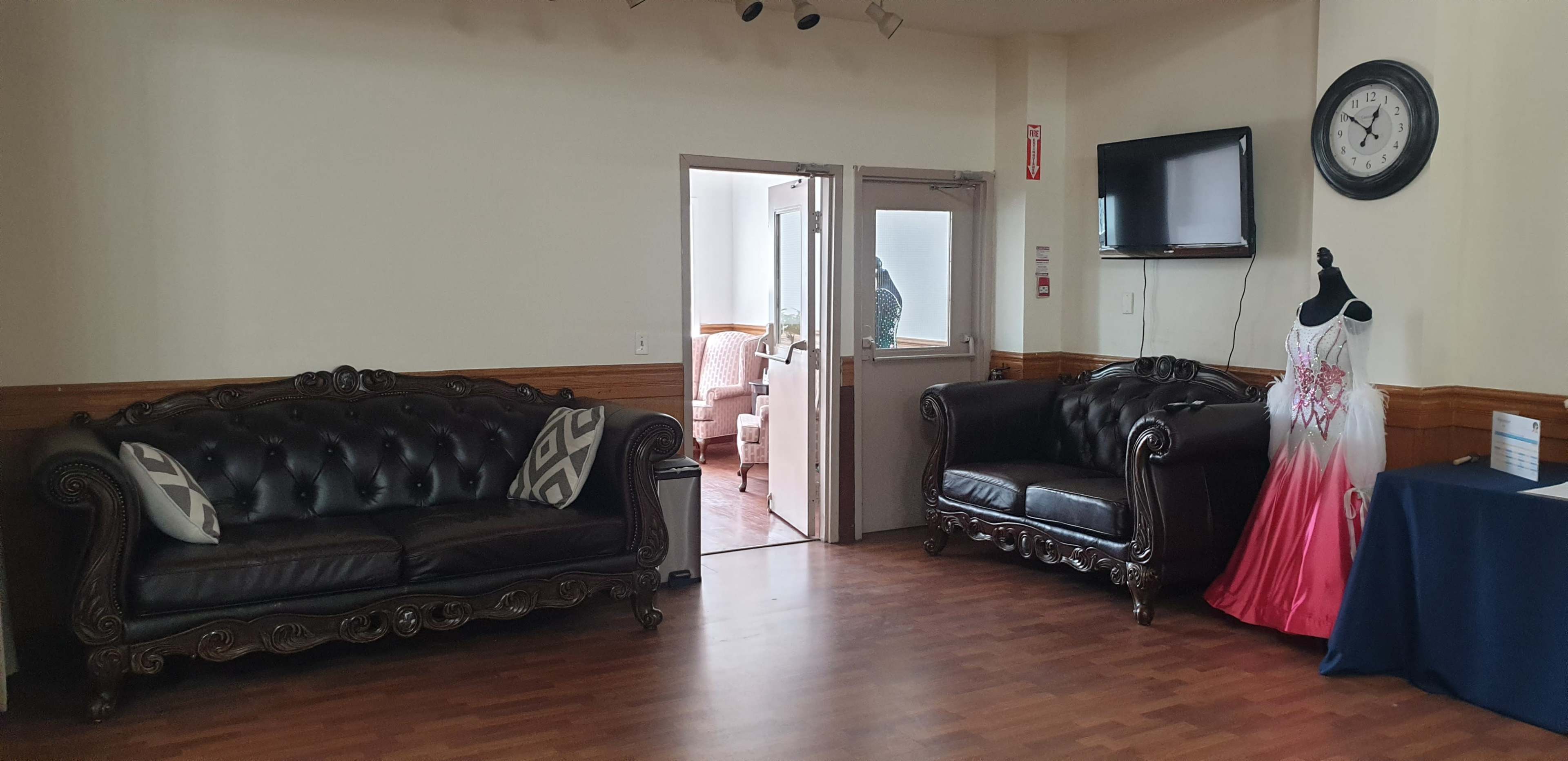 The scene features two black leather sofas, a doorway leading to another room, a wall clock, and a formal dress displayed on a mannequin.