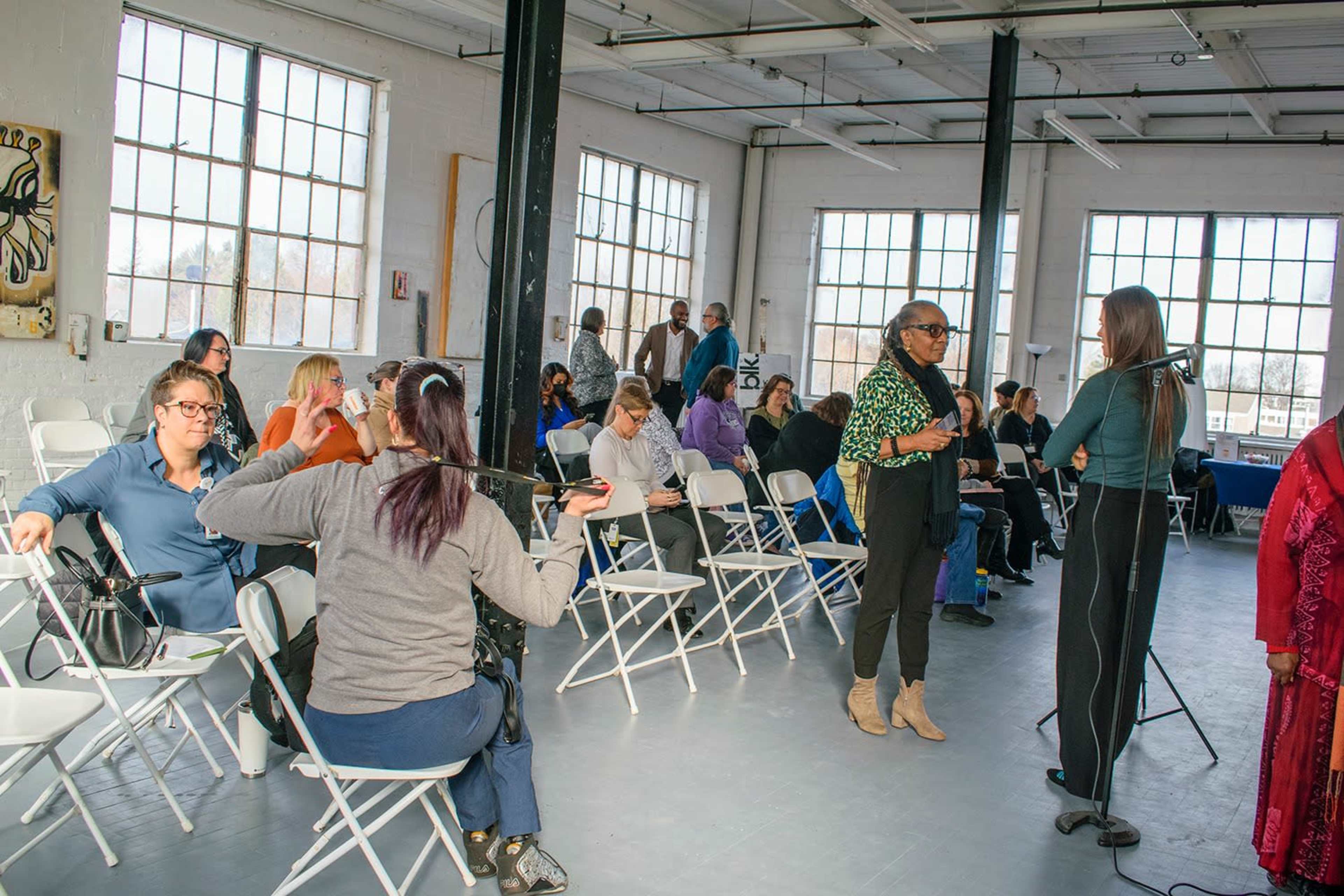 A group of people are gathered in a spacious room with large windows, engaged in conversation and preparing for an event.