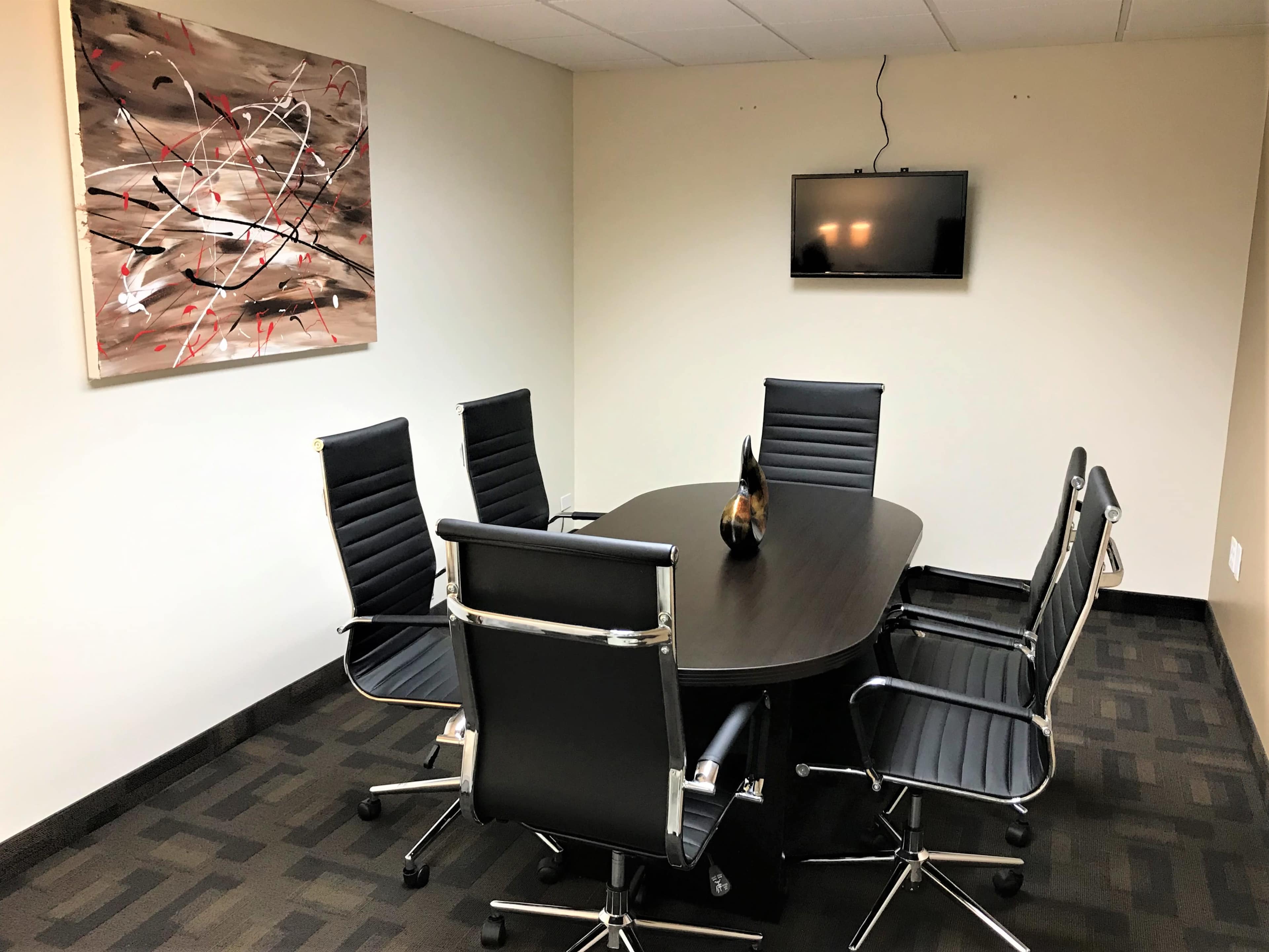 A conference room features a large oval table surrounded by six black leather chairs, with an abstract painting on the wall and a television mounted above.
