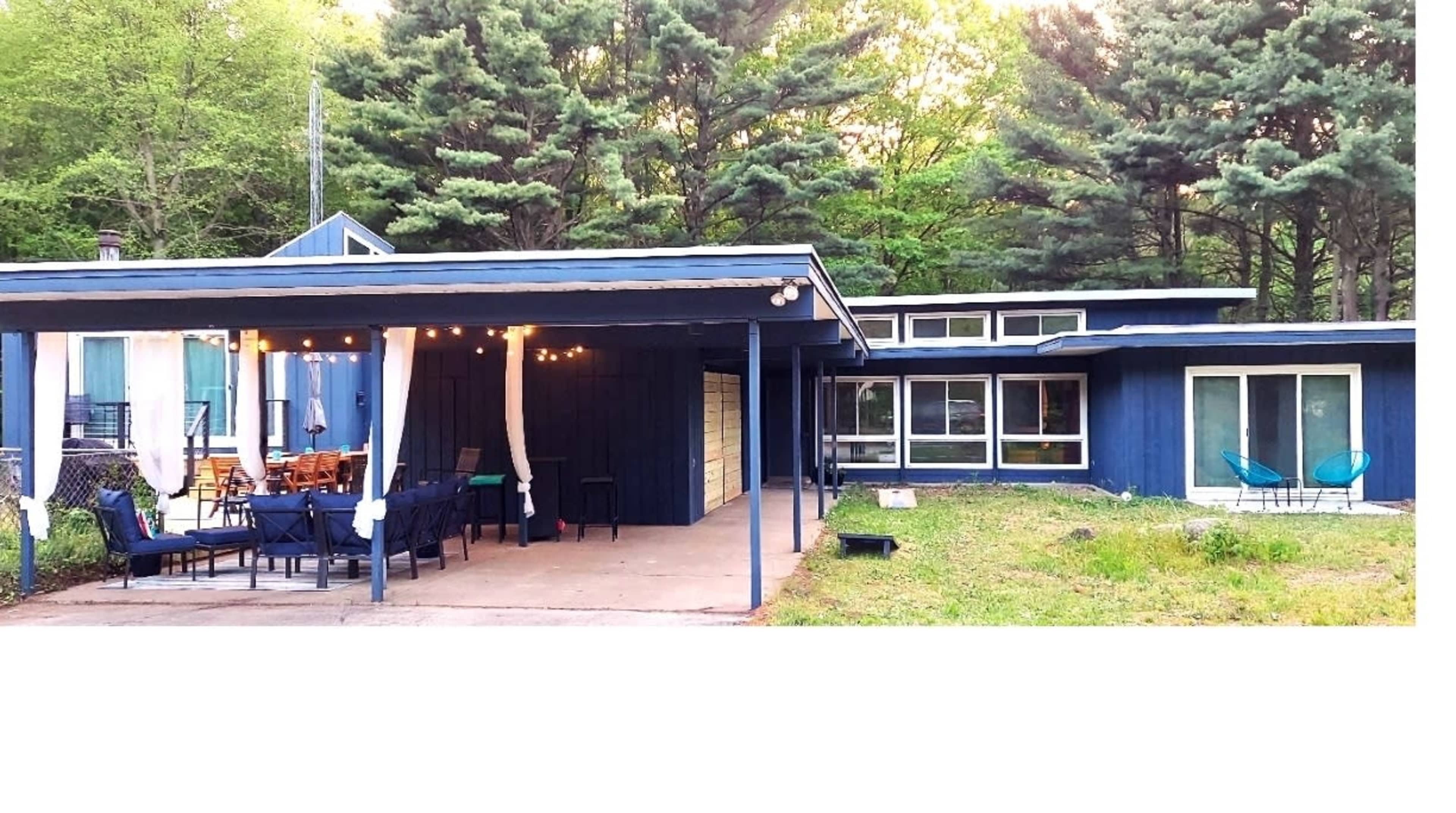 A blue and white cabin features a covered patio with string lights, surrounded by tall trees.
