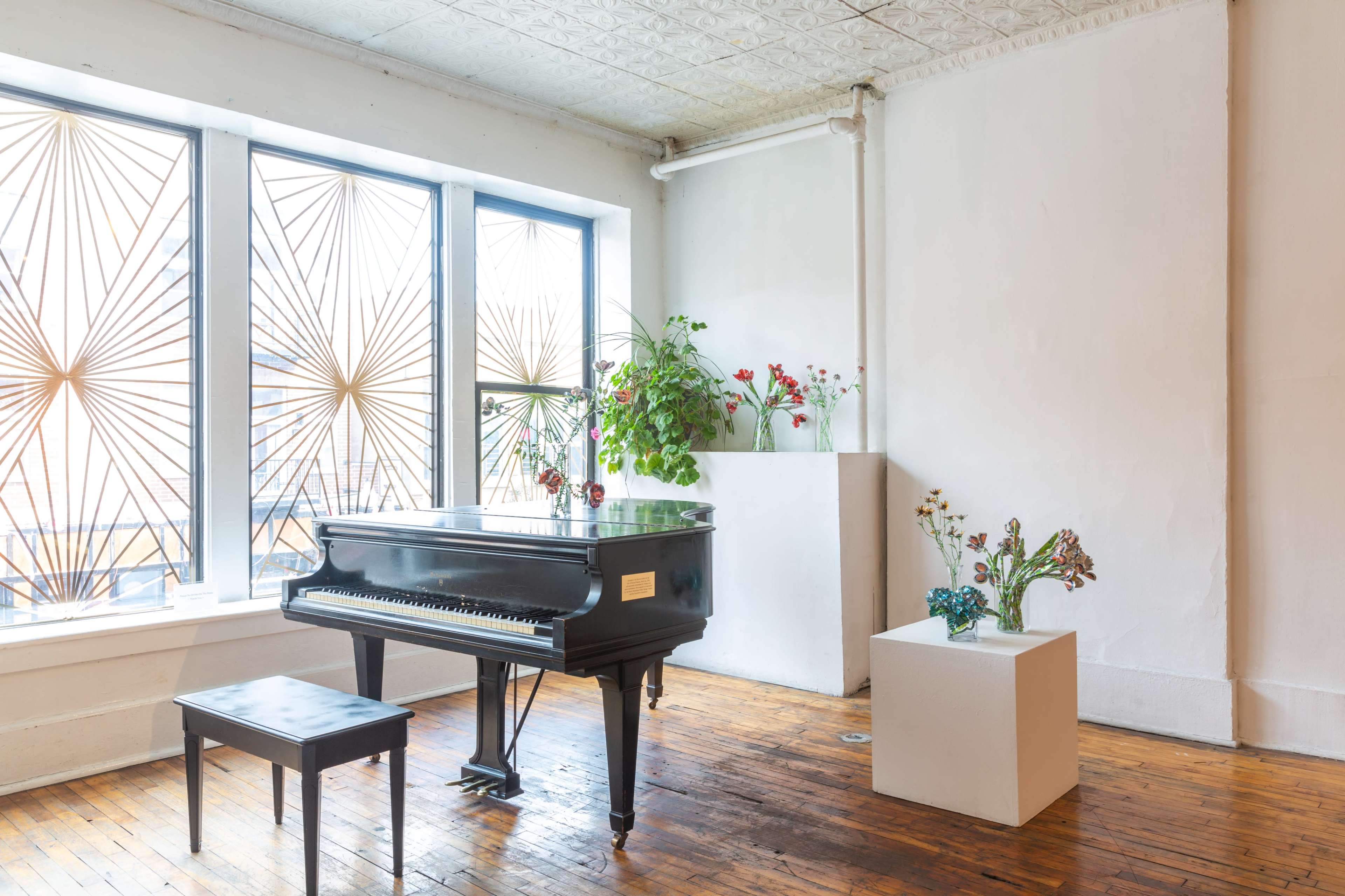 A black grand piano sits next to a small bench in a well-lit room with large windows and a white pedestal displaying floral arrangements.
