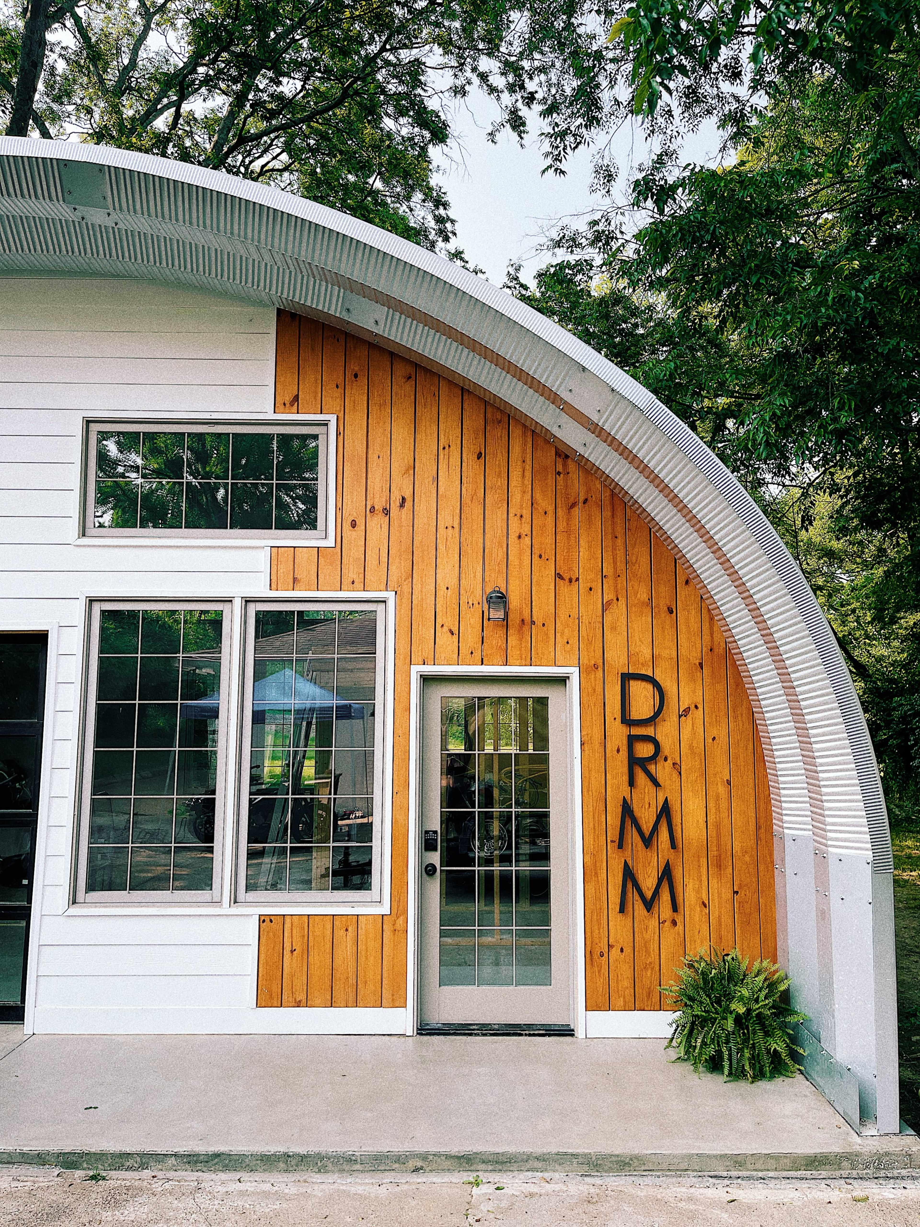 A modern, curved building with a combination of white siding and wooden panels, featuring large windows and the letters "DRMM" prominently displayed on the front.