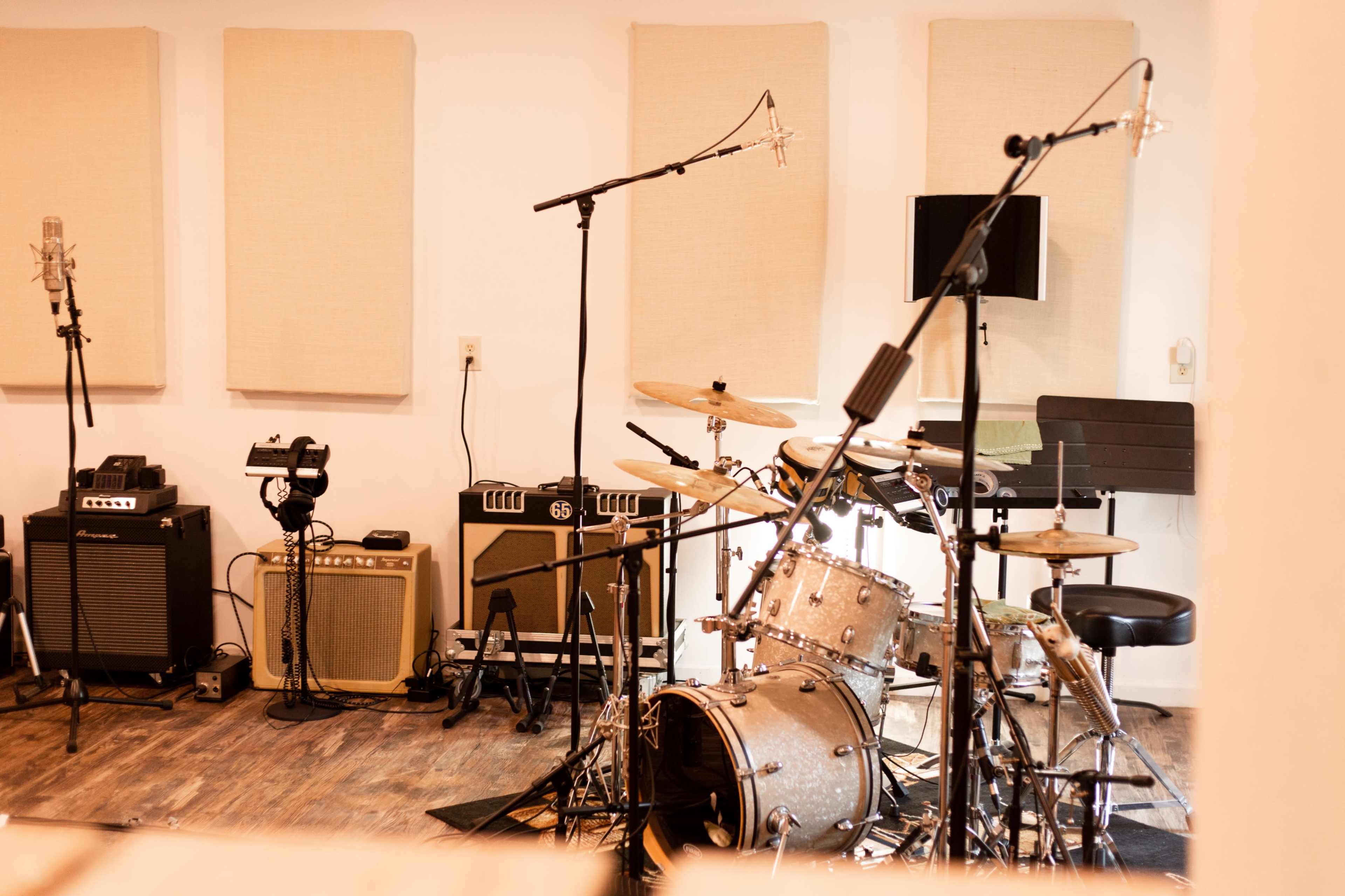 A music studio is equipped with microphones, amplifiers, and a drum set in front of soundproof panels.
