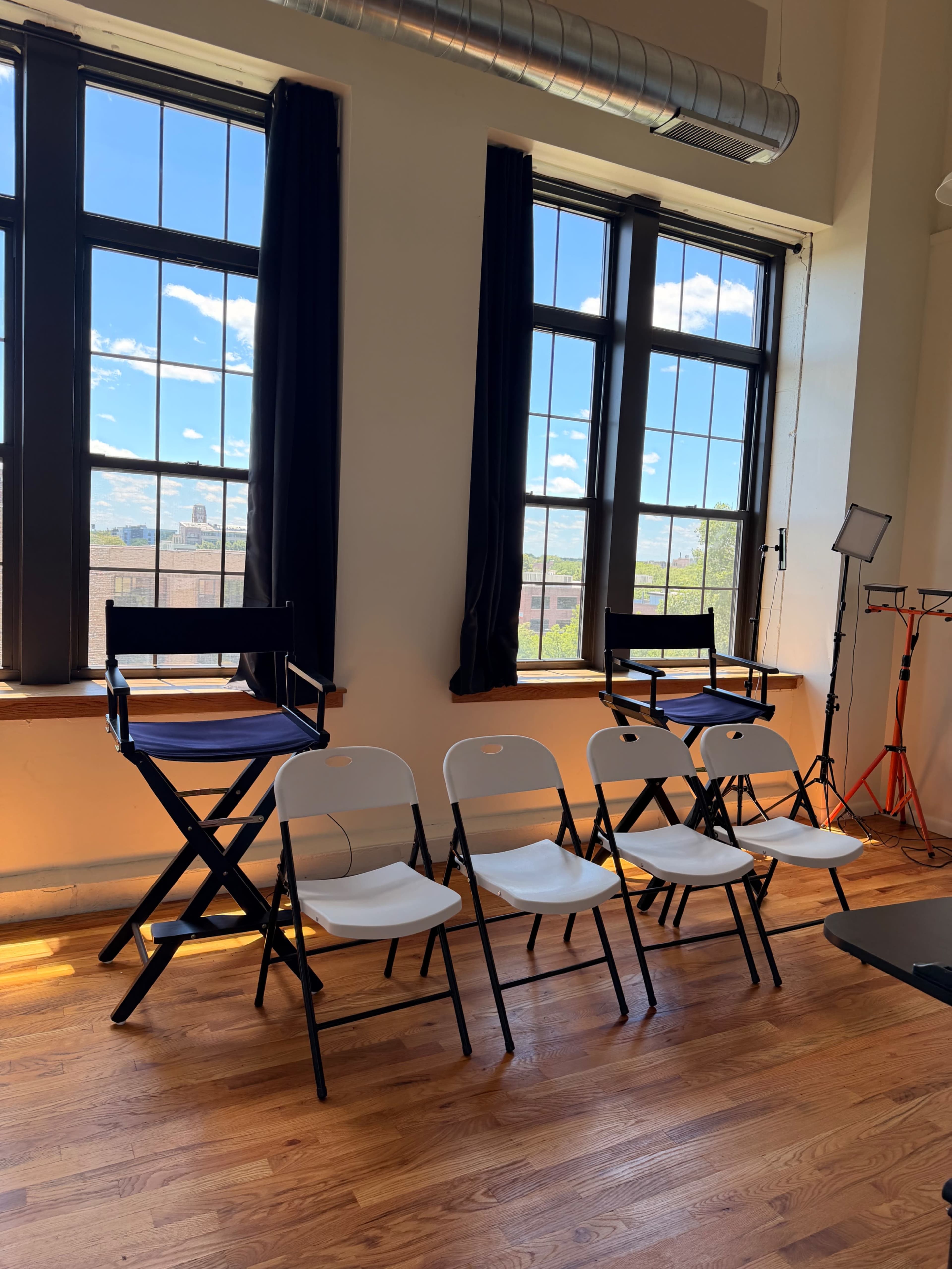The image shows a brightly lit room with large windows, featuring empty folding chairs arranged in a row and a director's chair in the foreground.