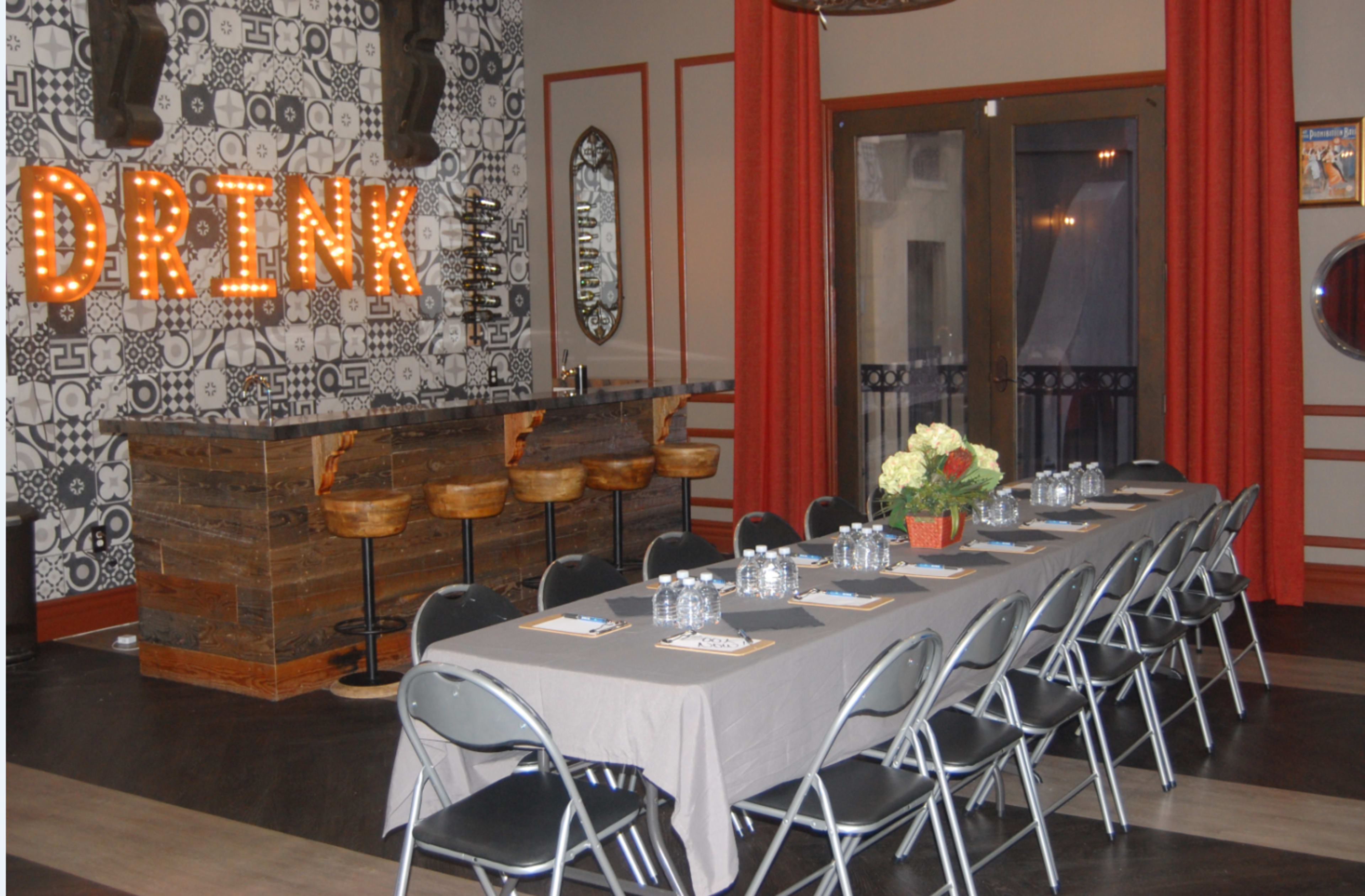 A long table with gray tablecloths is set up in a room featuring a bar with illuminated letters spelling "DRINK" and red curtains.