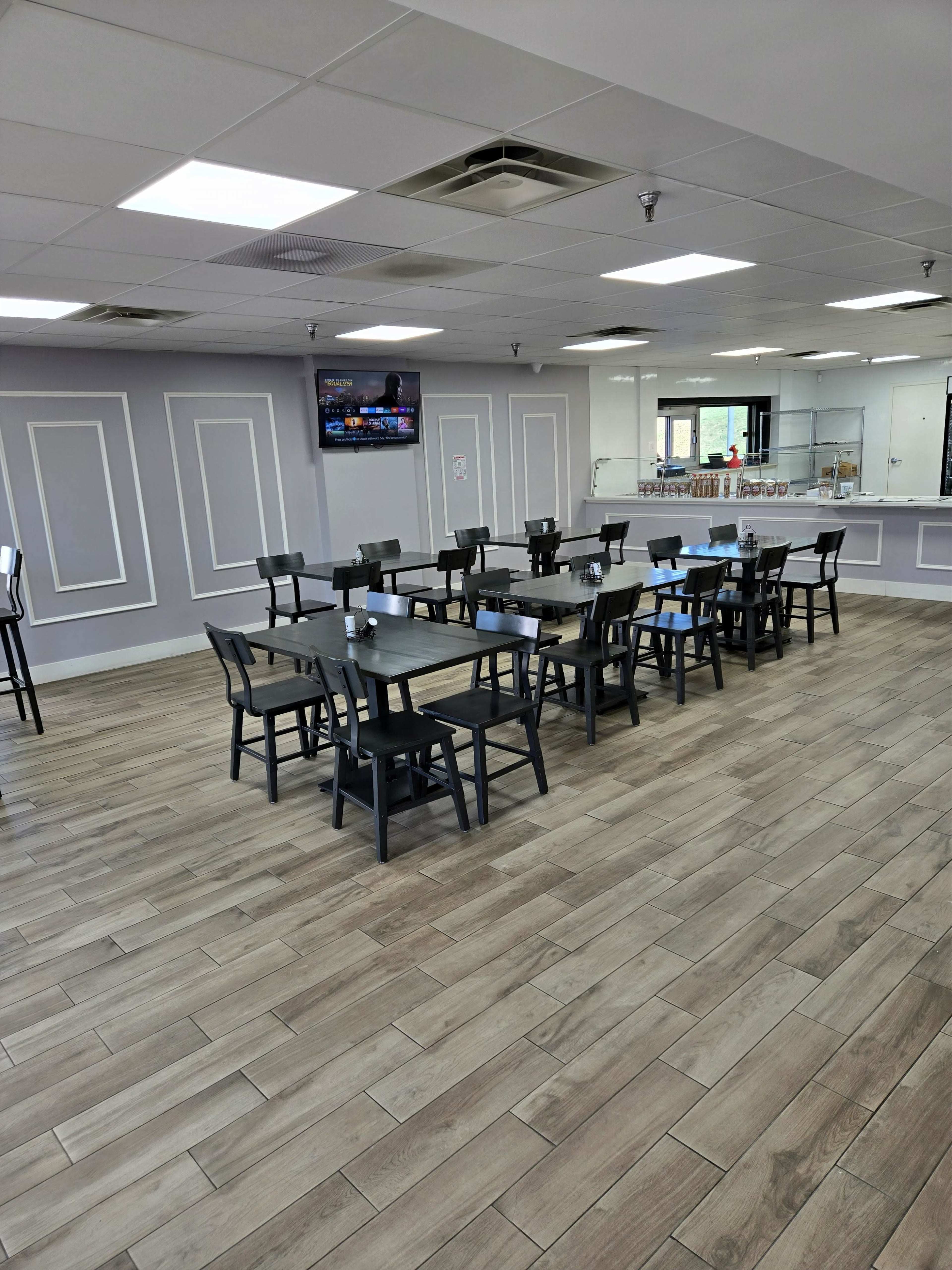 The image shows a modern dining area with wooden floors, black chairs, and tables arranged in rows, complemented by a television mounted on the wall.