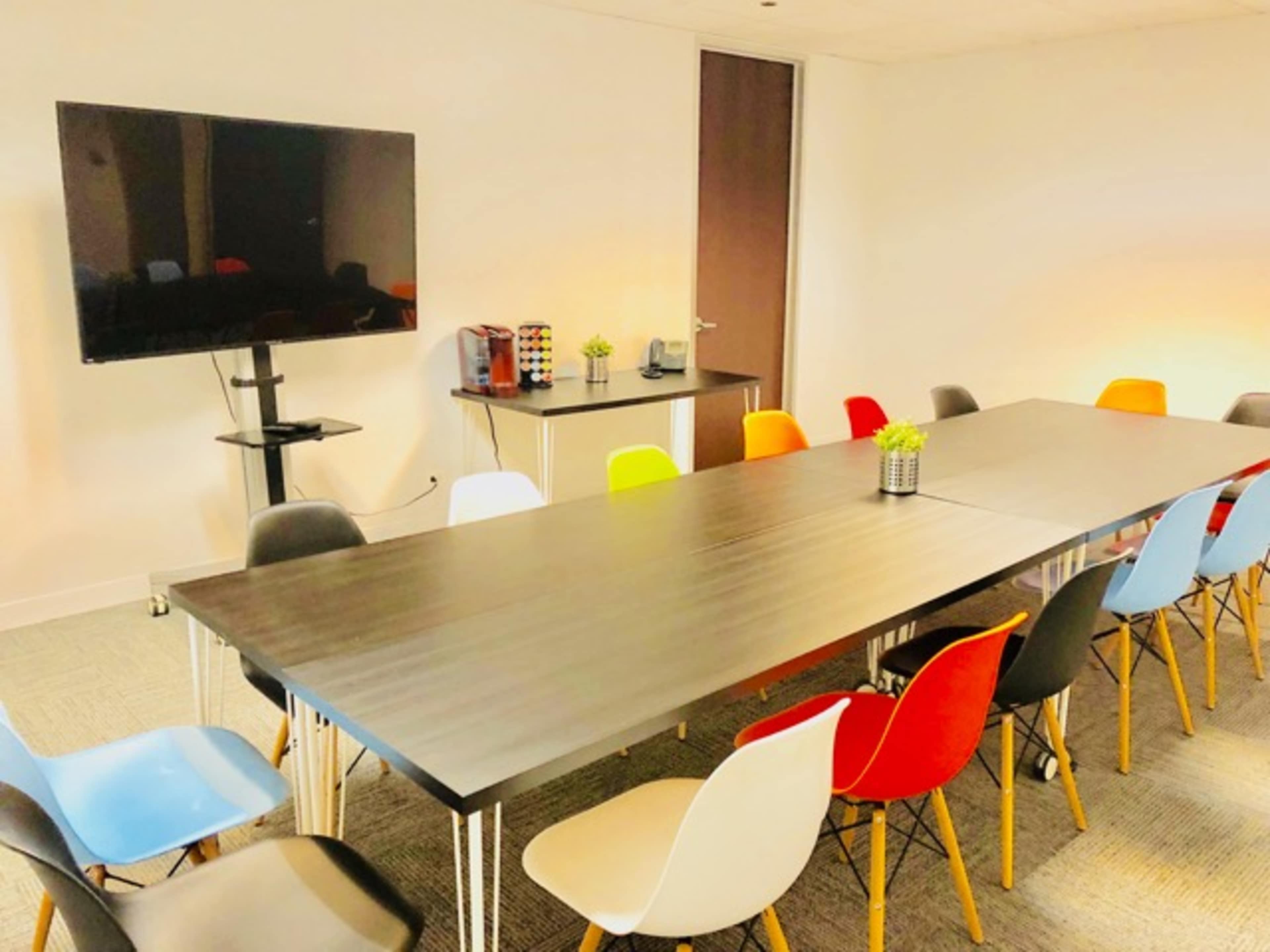 A modern conference room features a long table surrounded by colorful chairs and a large television mounted on the wall.